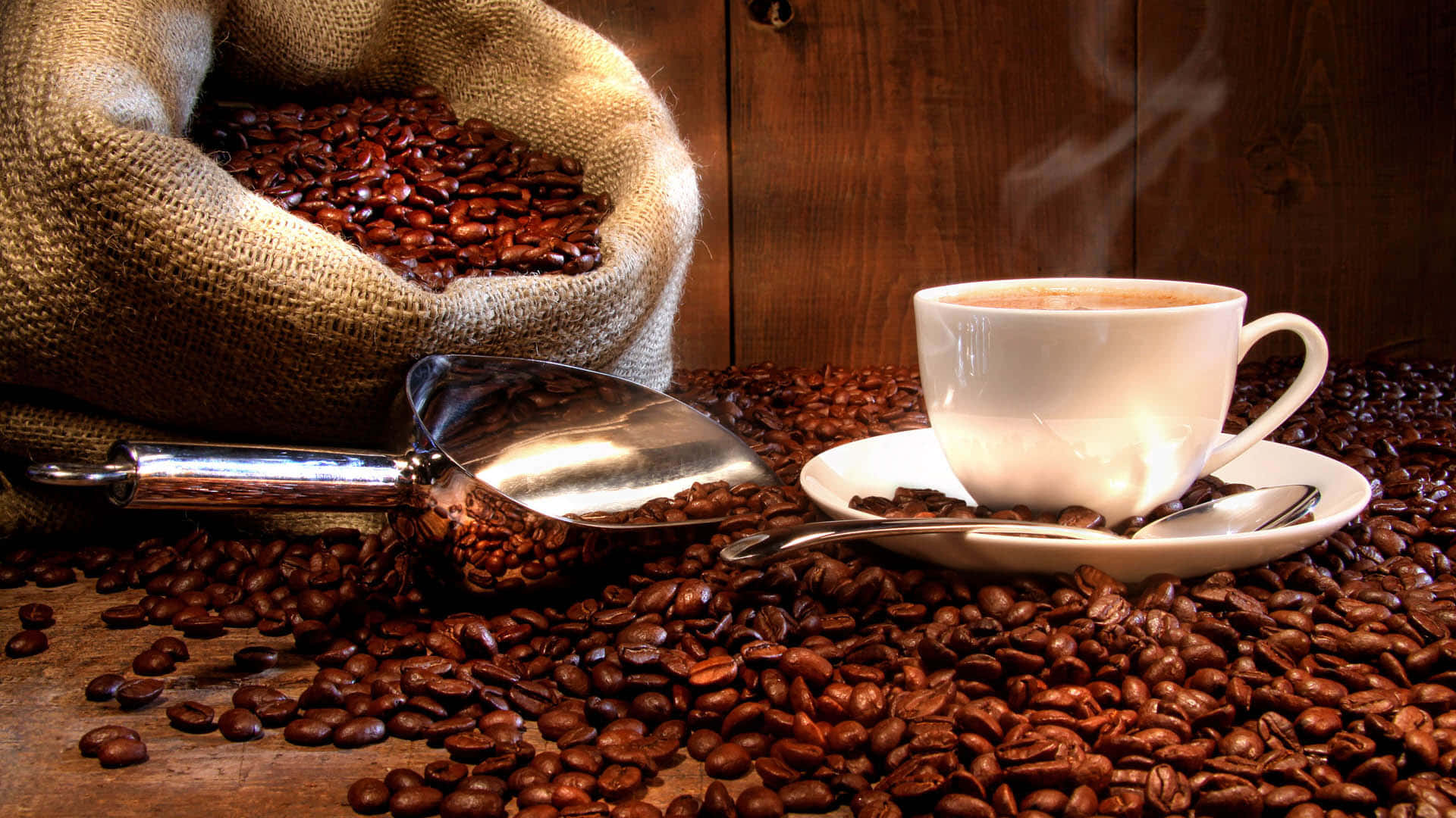 Brown Coffee 桌布 壁紙 — 一個冒著蒸氣的白咖啡杯放在碟子上，周圍是烘焙咖啡豆，背景有一個滿溢的麻布袋。