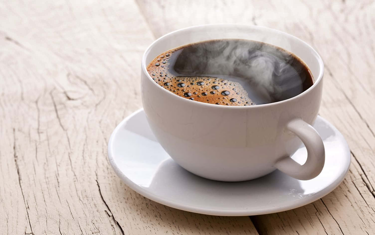 Coffee Cup 桌布 壁紙 — 一個冒著蒸氣的白色咖啡杯，裝著濃郁的深色咖啡和泡沫奶泡，放在碟子上，置於一張經風化的淺色木桌上。