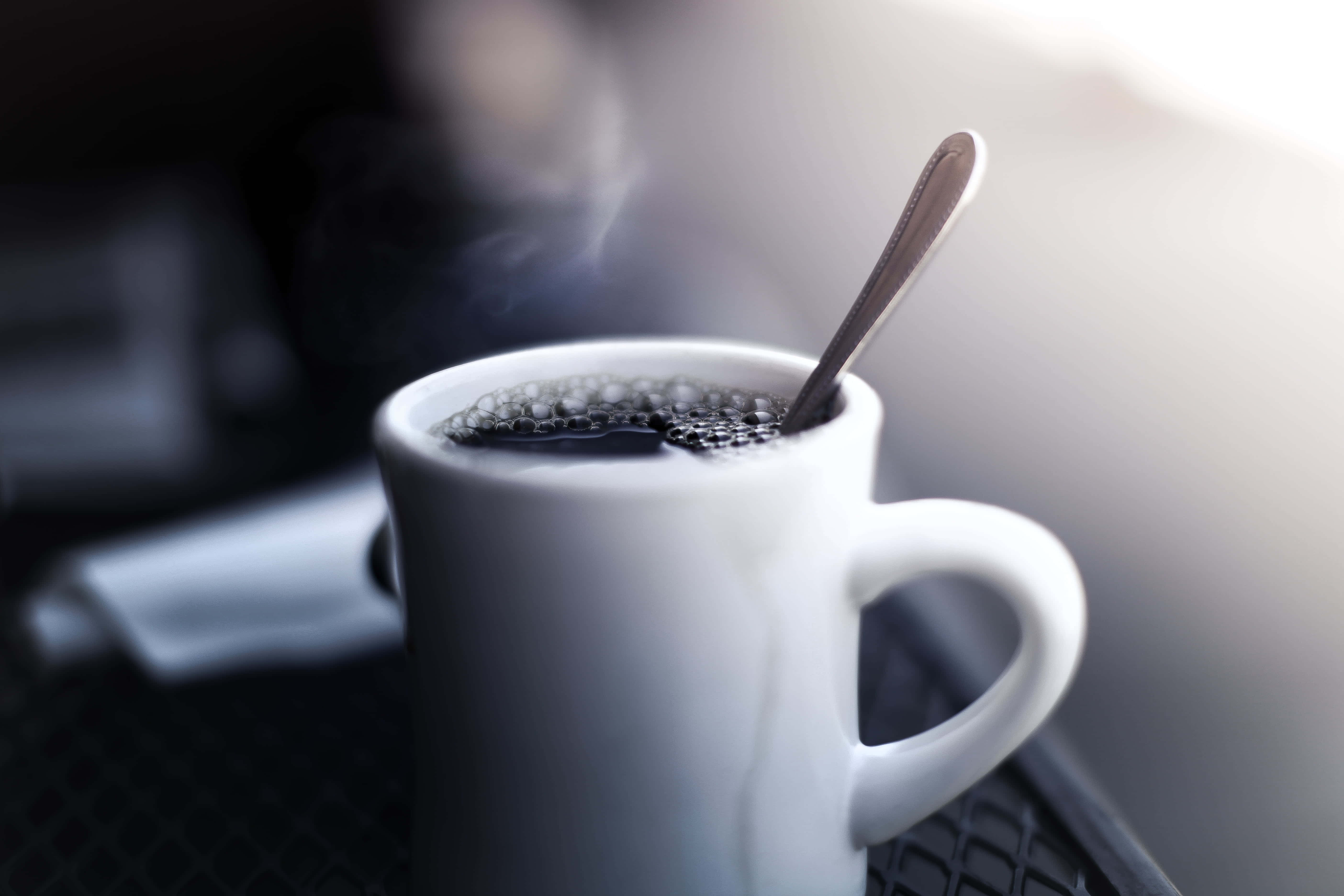 Black Coffee 桌布 壁紙 — 一個冒著蒸氣的白色馬克杯，裝著黑咖啡，附有湯匙，放在有紋理的表面上，沐浴在柔和背光中。