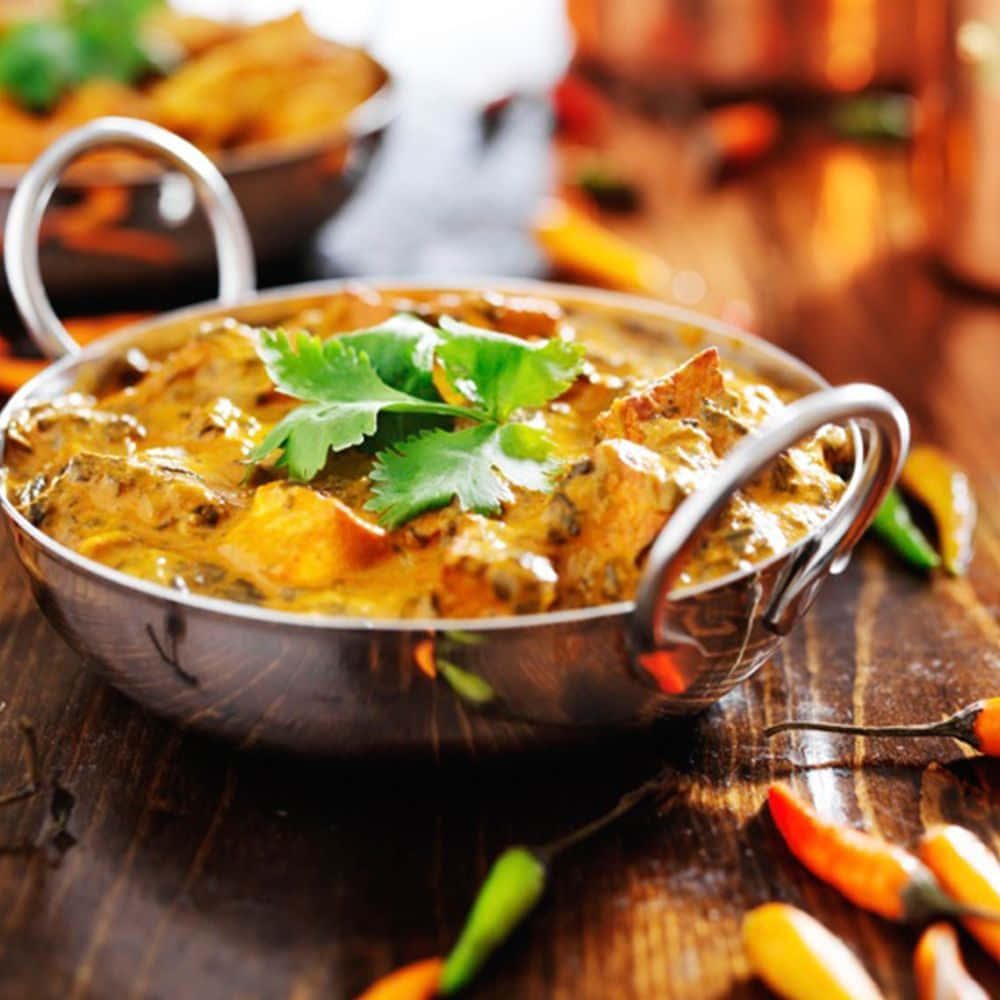 Indian Food 桌布 壁紙 — 一個冒著蒸氣的金屬碗，盛滿濃郁的黃咖喱雞和新鮮香菜，放在質樸的木桌上，周圍環繞著色彩繽紛的辣椒。