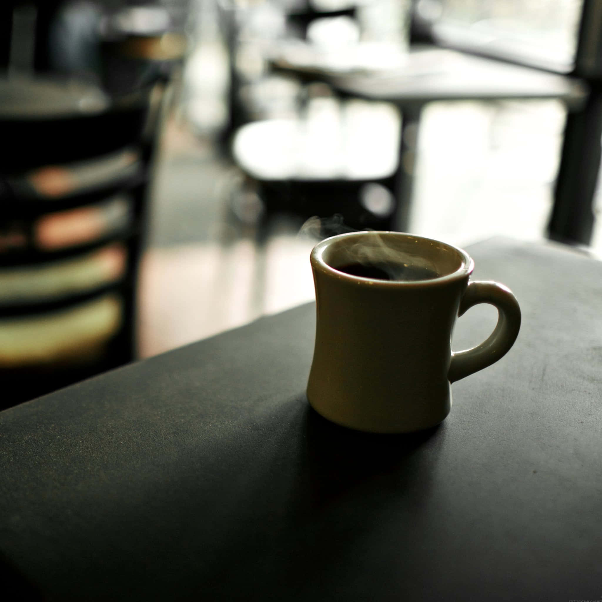 Black Coffee 桌布 壁紙 — 一個冒著蒸氣的陶瓷咖啡杯放在昏暗的桌子上，背景是燈光柔和、有模糊椅子和窗戶的咖啡館。
