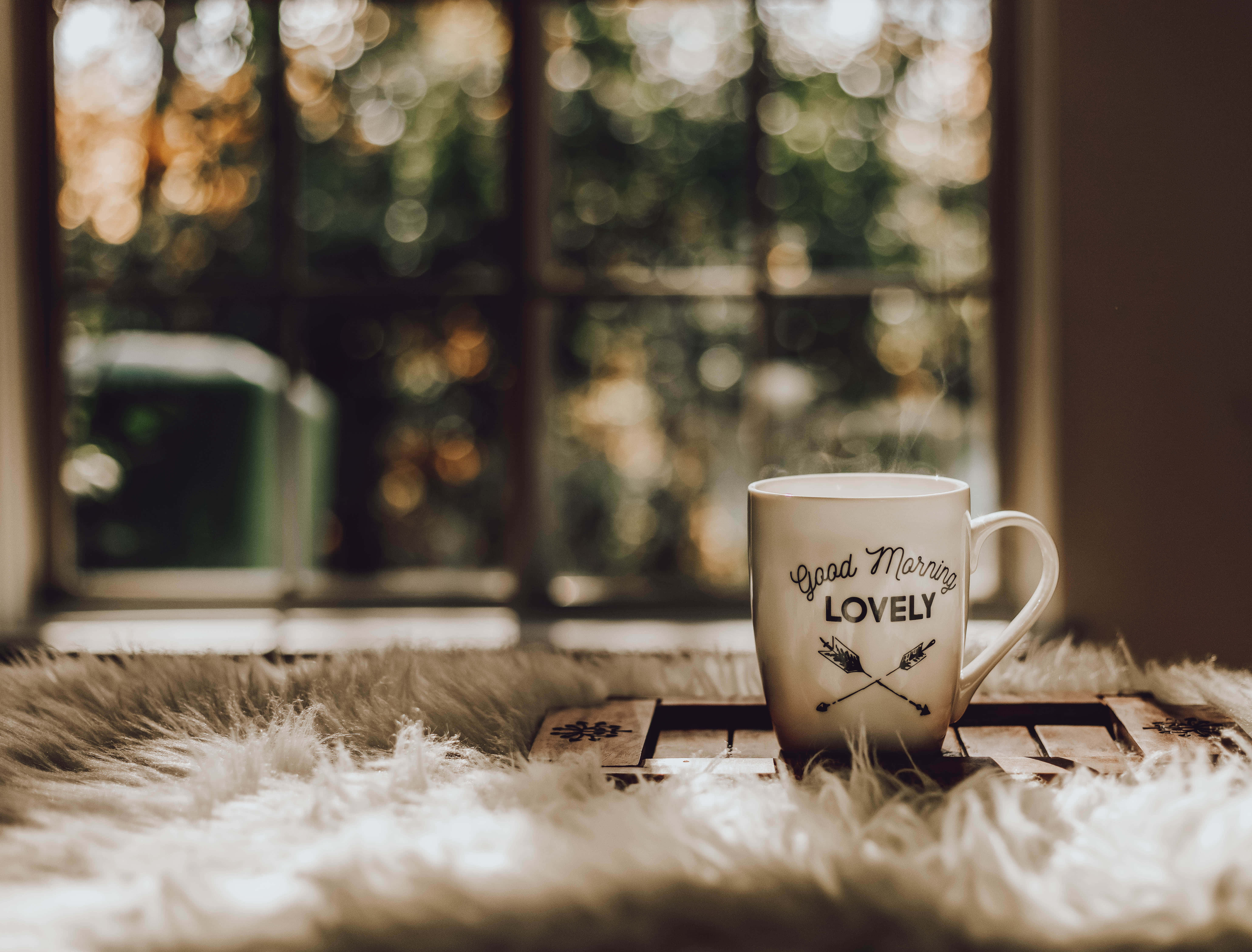 Brown Coffee 桌布 壁紙 — 一個冒著蒸氣的馬克杯，上面寫著「Good Morning LOVELY」，放在陽光照射的窗邊一個毛絨白色地毯上的木托盤上。