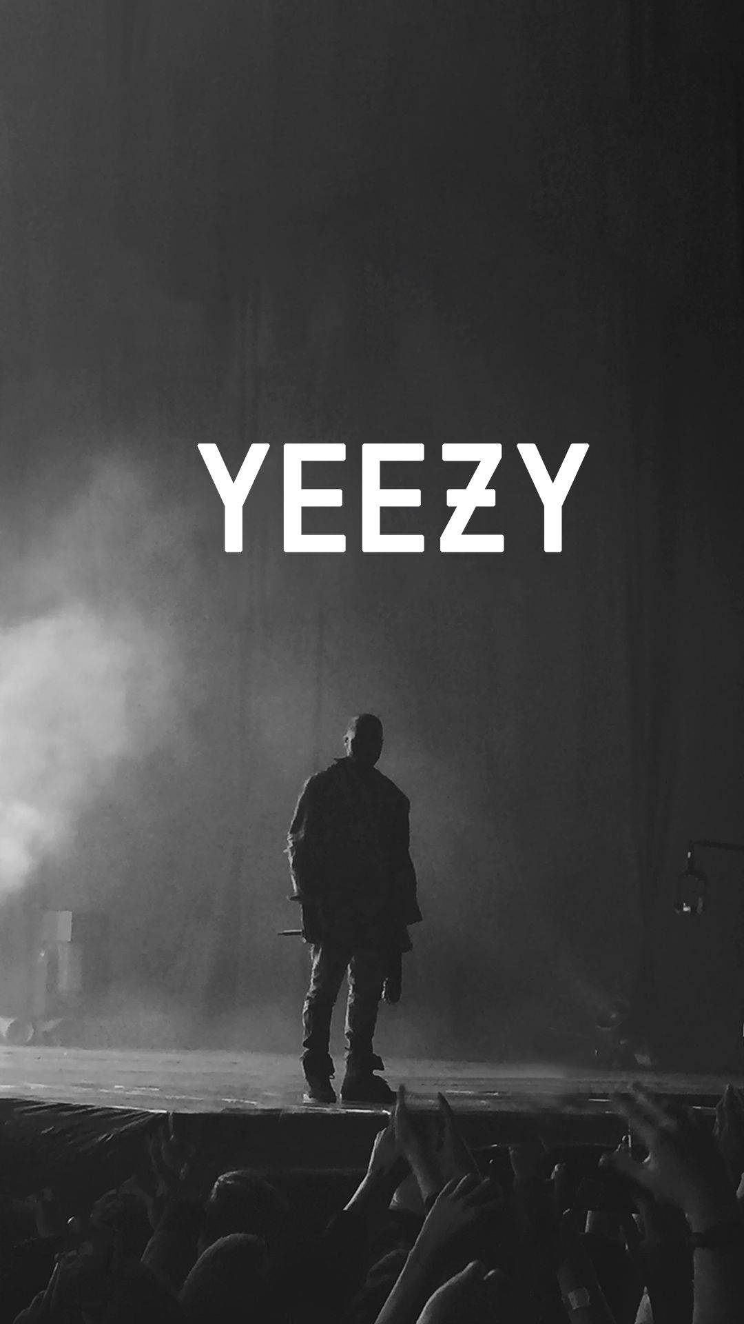 Kanye West Yeezy In White 壁紙 — 一個剪影人物站在煙霧彌漫的舞台上，'YEEZY' 文字下方，人群的手在黑暗中向上伸出。