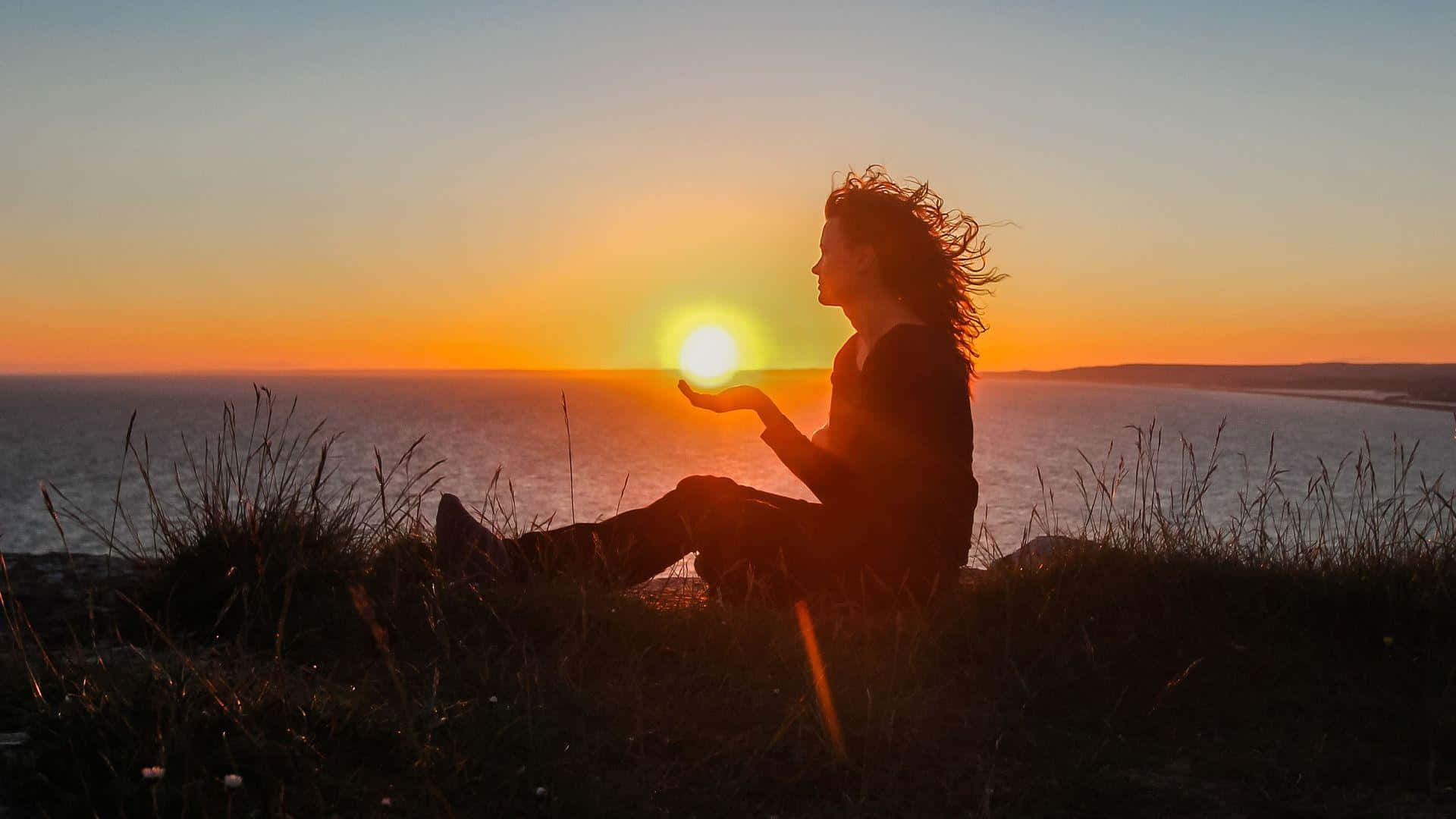 Sunset Silhouette 桌布 壁紙 — 一個剪影女子坐在懸崖邊，日落時分，她的手捧著海面上發光的太陽。