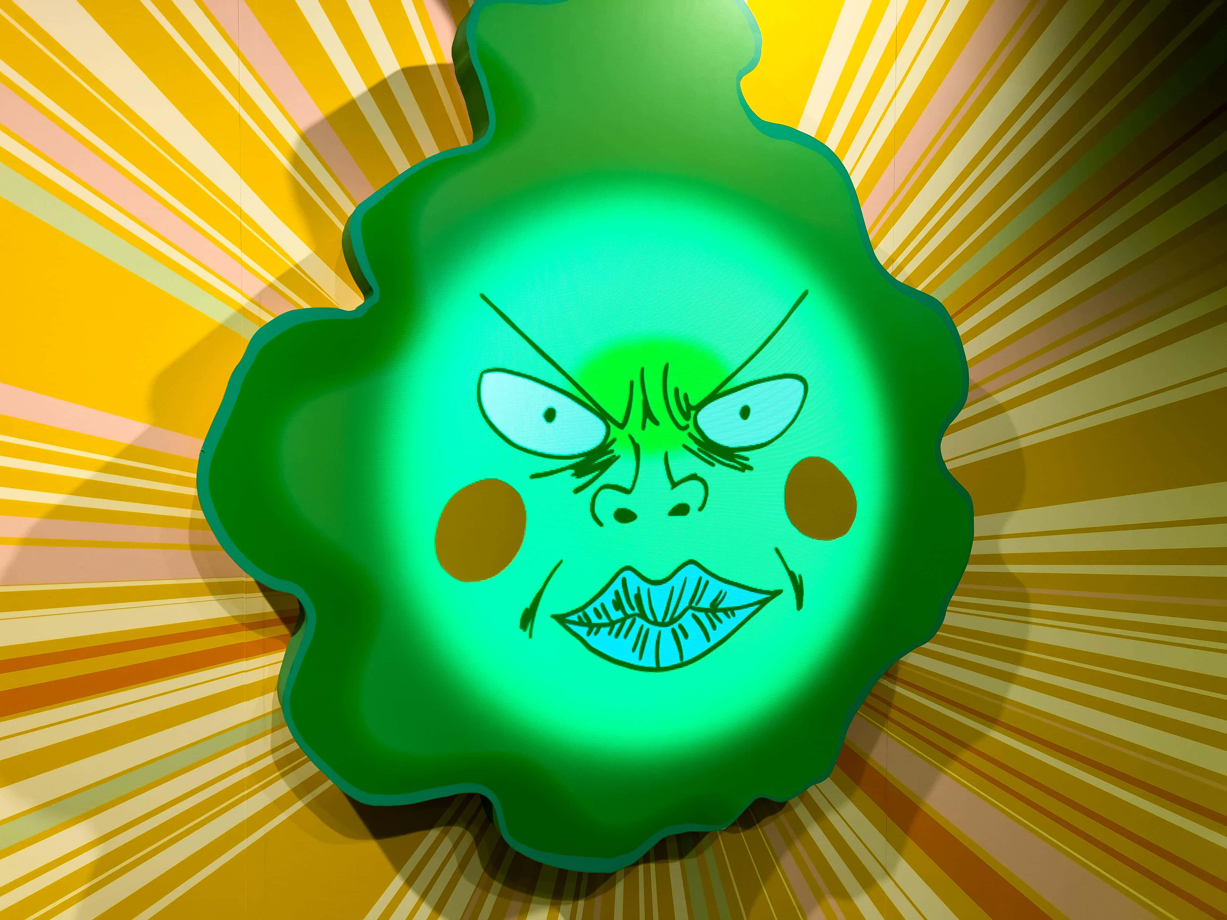 Mob Psycho 100 Ekubo 桌布 壁紙 — 一個卡通風格的綠色憤怒面孔，帶有發光的眼睛和臉頰，在陽光四射的黃色背景上閃耀。