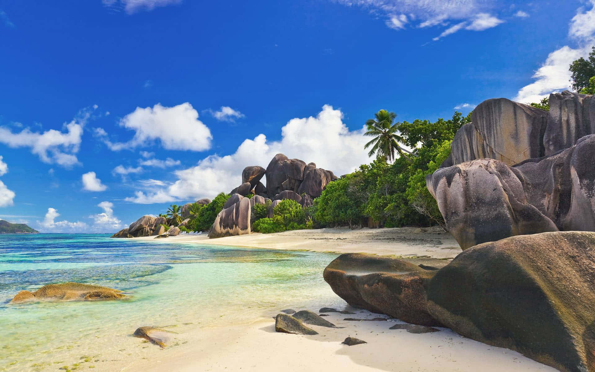 Seychelles Beach 桌布 壁紙 — 一個原始熱帶海灘，擁有碧綠海水、白色沙灘和巨大花崗岩巨石，在明亮的藍天和蓬鬆的白雲下。
