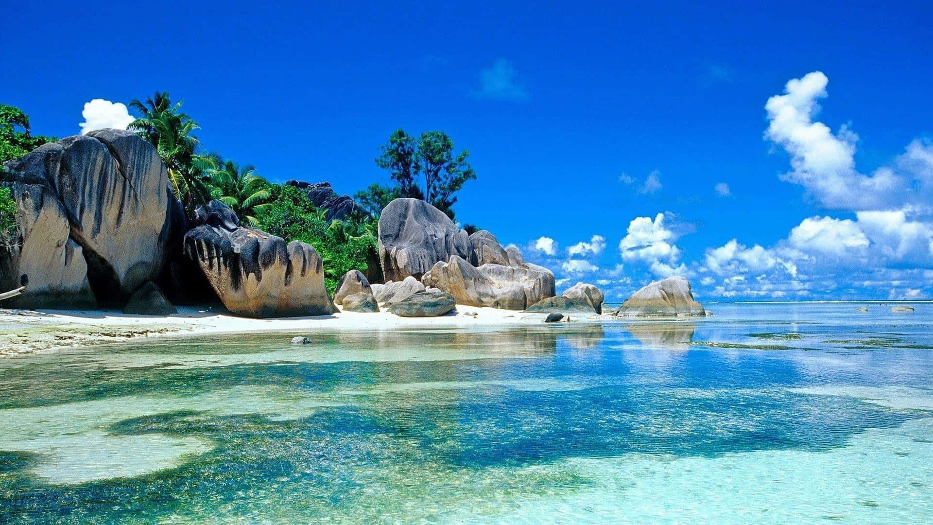 Beach Desktop 桌布 壁紙 — 一個原始熱帶海灘，擁有碧綠海水和巨大岩石，在明亮的藍天和蓬鬆的白雲下。