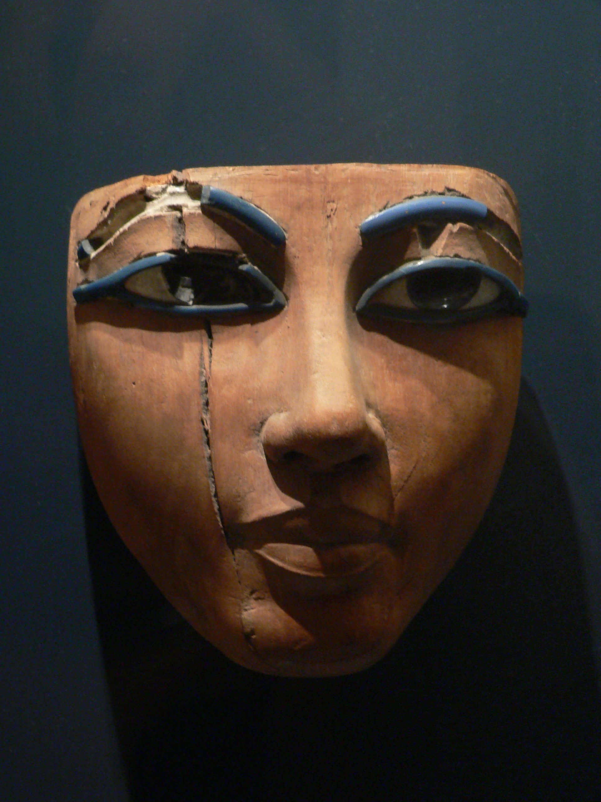 Ancient Egypt 桌布 壁紙 — 一個古埃及木製死亡面具，帶有藍色塗繪的眼睛和裂紋，在深色背景下展示。