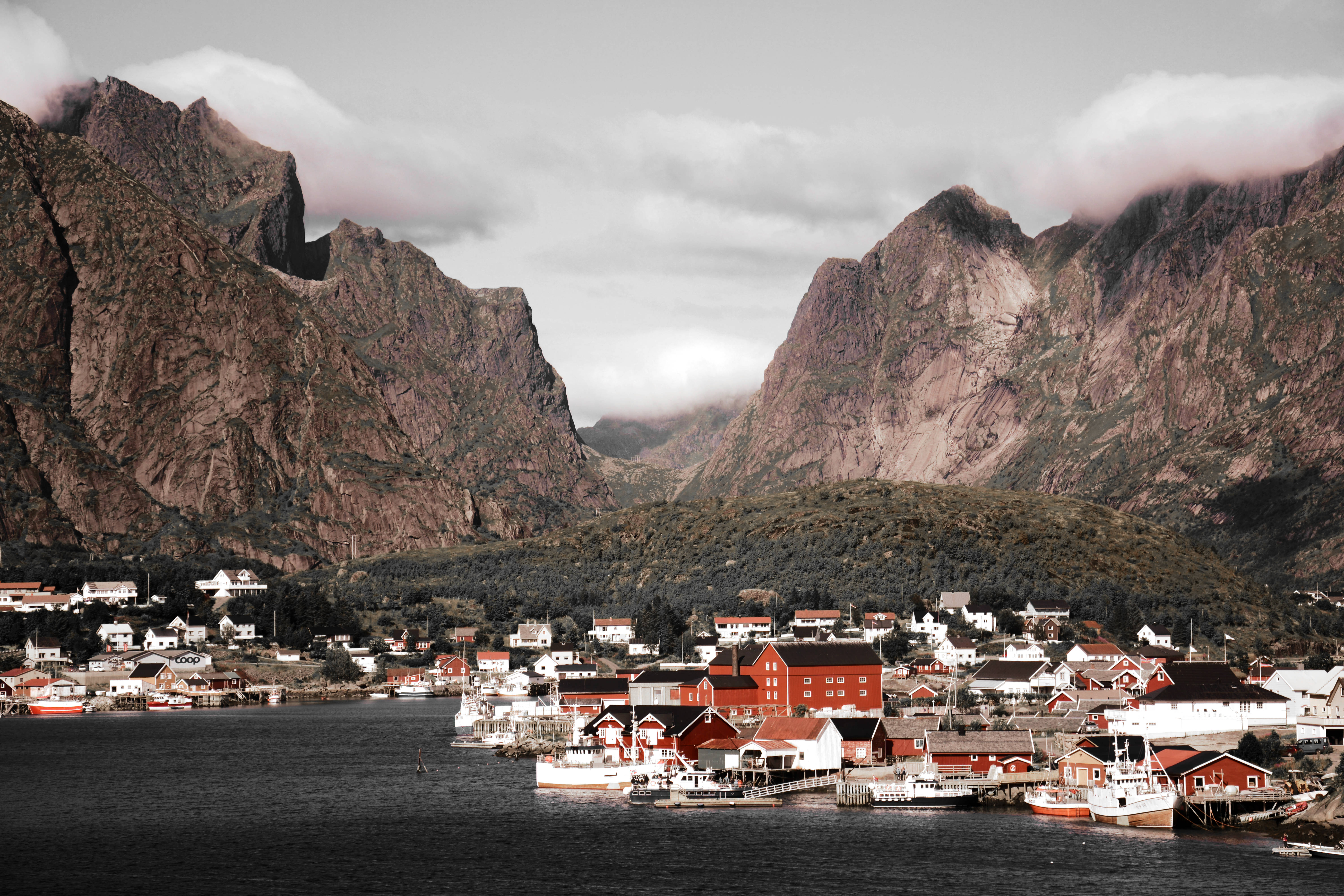 Norway 桌布 壁紙 — 一個古樸的海岸村莊，紅瓦屋頂的房屋坐落在高聳的岩石山之間，天空多雲。