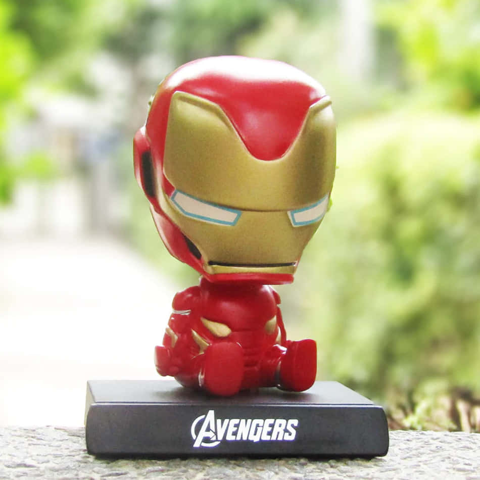 Iron Man Bobbleheads 桌布 壁紙 — 一個可愛的Q版鐵甲奇俠公仔坐在戶外的復仇者聯盟基地上，背後有模糊的綠色樹葉。