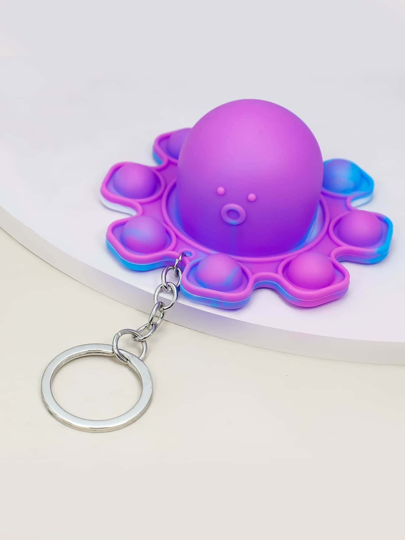 Popit 桌布 壁紙 — 一個可愛的紫色和藍色章魚形減壓玩具，帶有金屬鑰匙扣，放置在白色表面上。