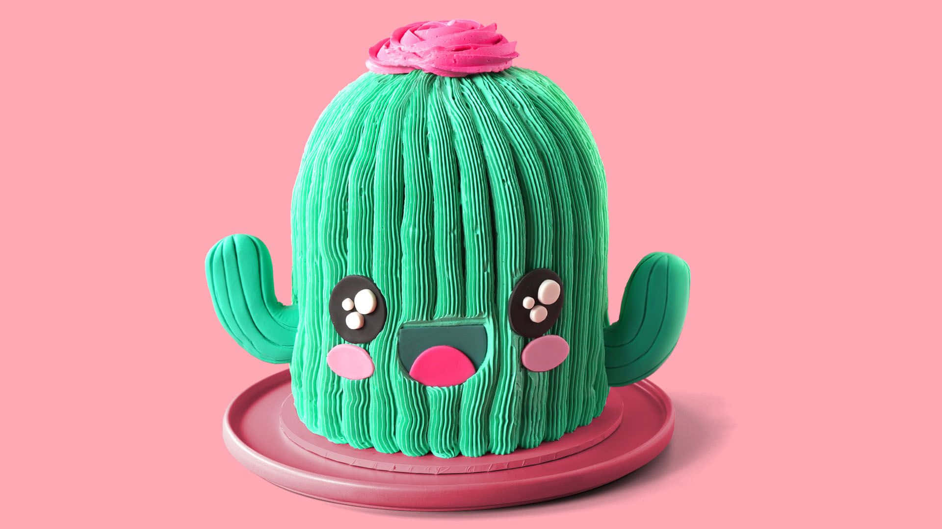 Kawaii Cactus 桌布 壁紙 — 一個可愛的綠色仙人掌蛋糕，帶有粉紅色糖霜和大眼睛，放在粉紅色碟子上，背景是柔和的粉紅色。