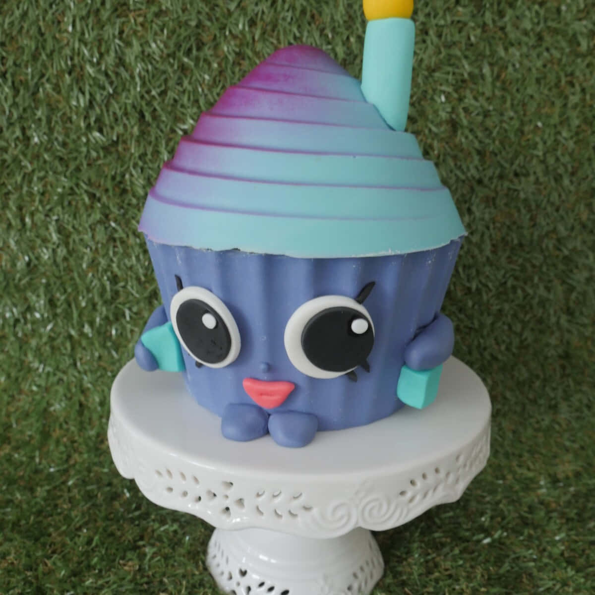 Shopkins 桌布 壁紙 — 一個可愛的藍色杯子蛋糕角色，有大眼睛和彩虹條紋帽子，坐在白色蛋糕架上，背景是綠色草地。