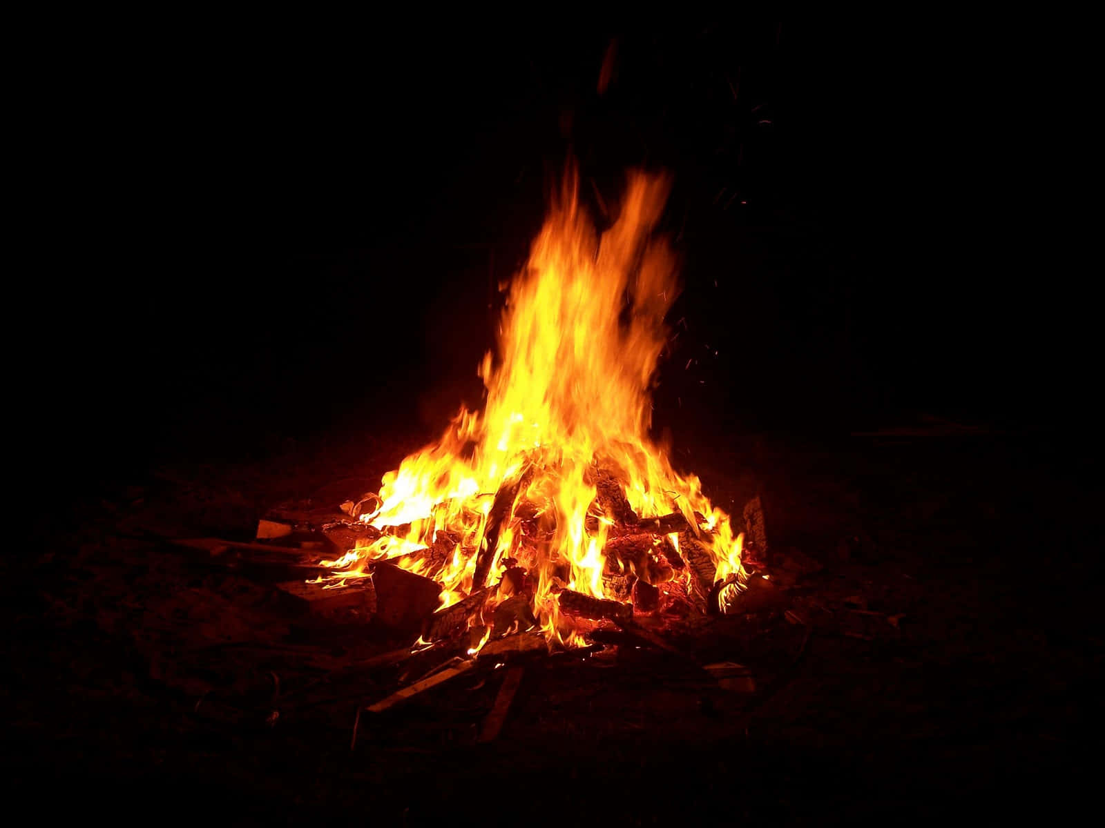 Fall Bonfire 桌布 壁紙 — 一個咆哮的營火在漆黑的夜空中明亮地燃燒，將溫暖的橙色光芒投射在周圍的木頭上。