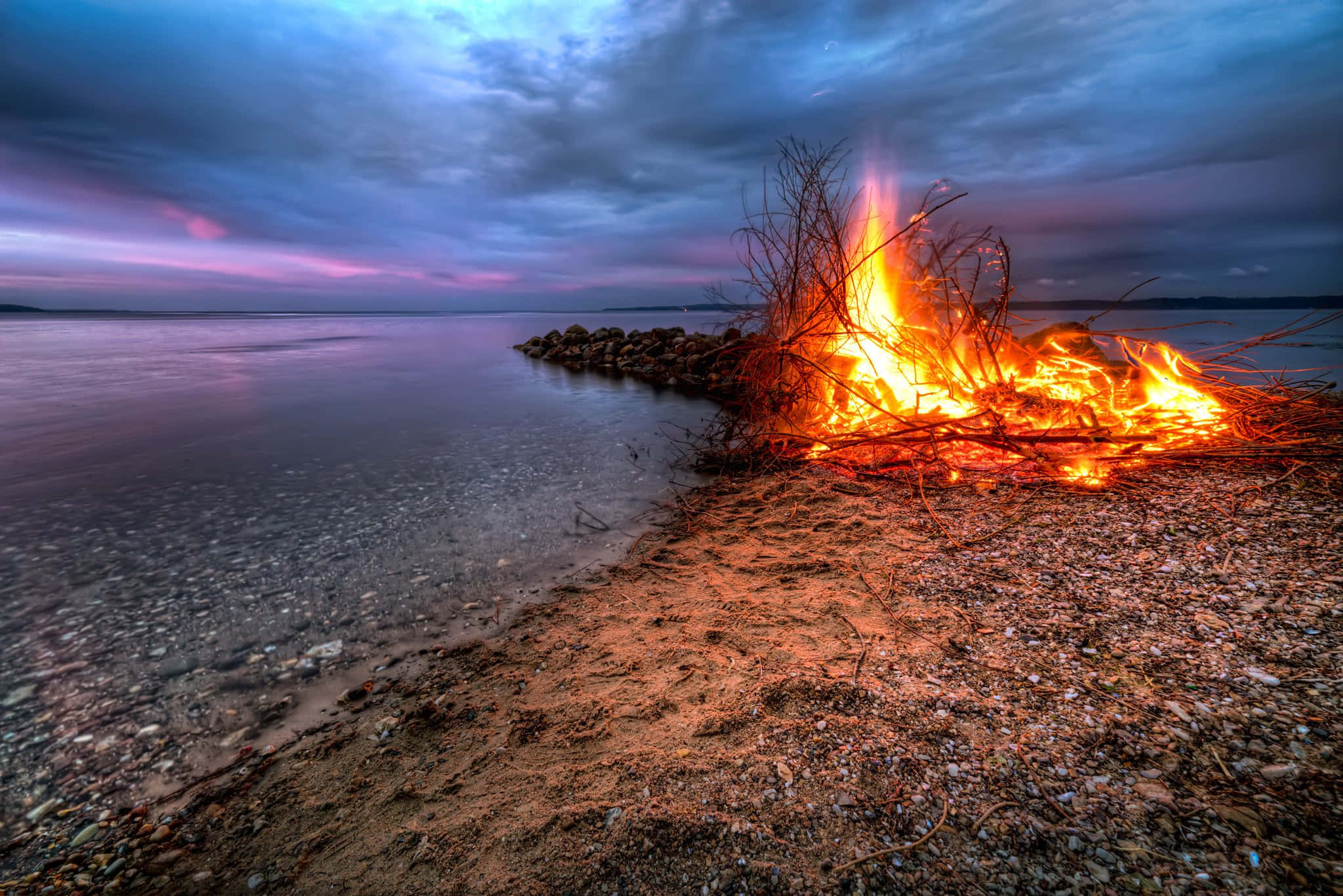 Fall Bonfire 桌布 壁紙 — 一個咆哮的營火在黃昏時分在卵石岸邊明亮地燃燒，向陰鬱的紫色和藍色天空投射溫暖的光芒。