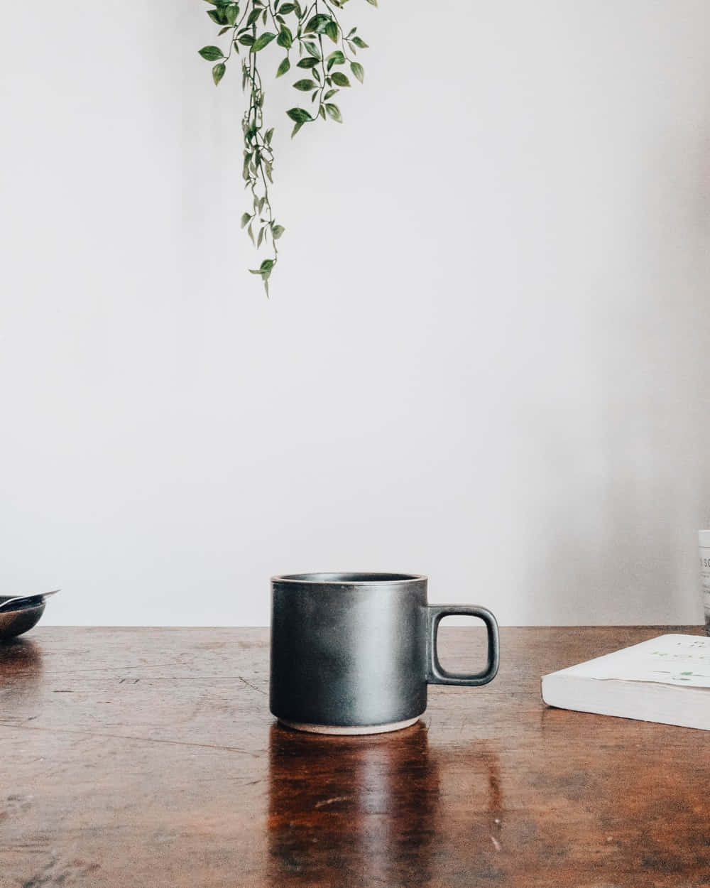 Minimalist Coffee 桌布 壁紙 — 一個啞光黑色馬克杯放在質樸木桌中央，旁邊有一本書和垂吊的綠色植物，背景是純白牆壁。