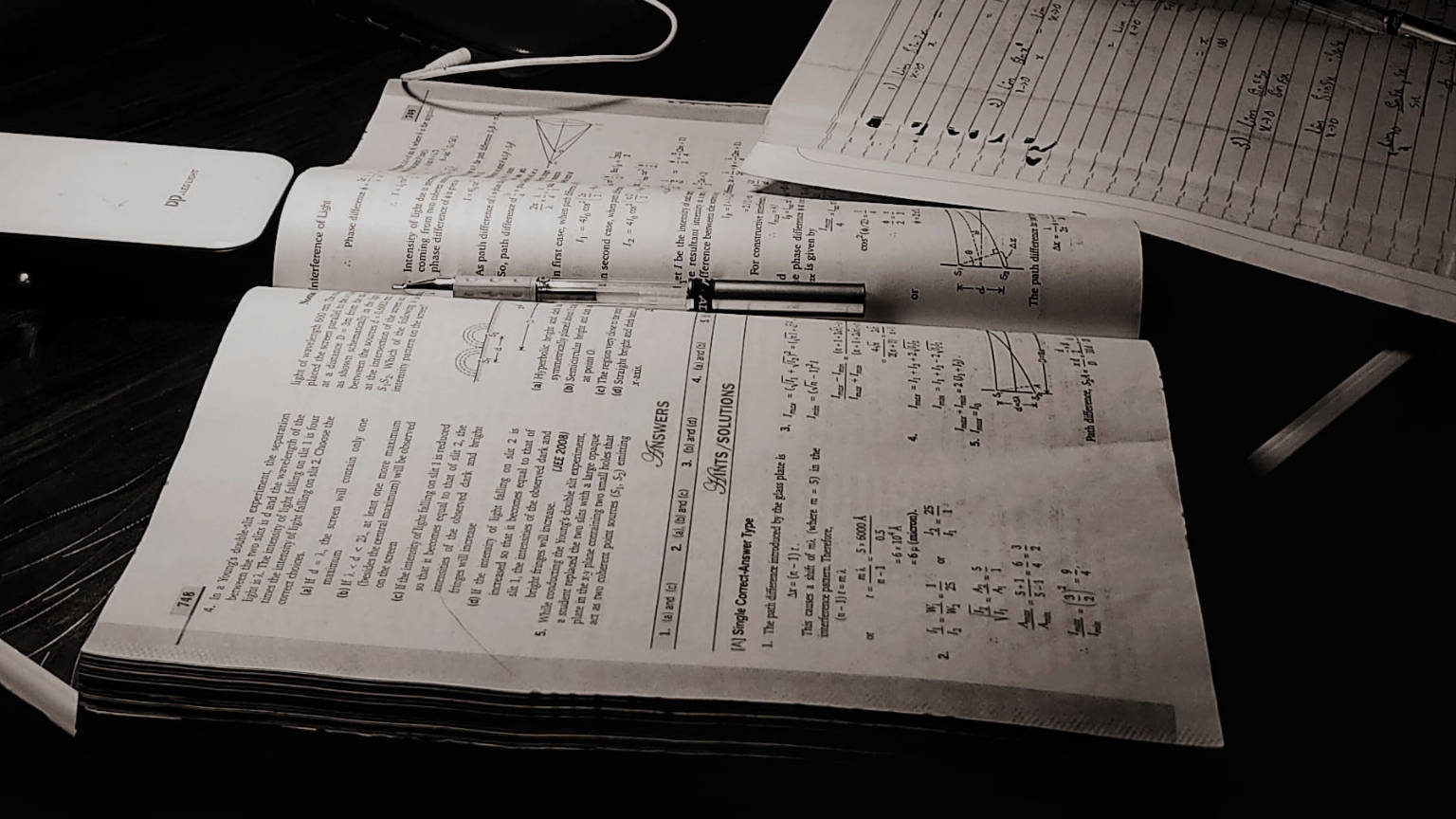 書籍 桌布 壁紙 — 一個單色特寫，顯示在深色木桌上打開的數學教科書和手寫解答，旁邊有筆和耳機。