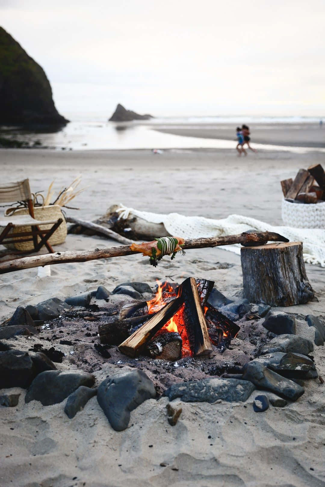 Winter Camping 桌布 壁紙 — 一個噼啪作響的海灘營火，串著烤肉的棍子，周圍是岩石和浮木，天空多雲。