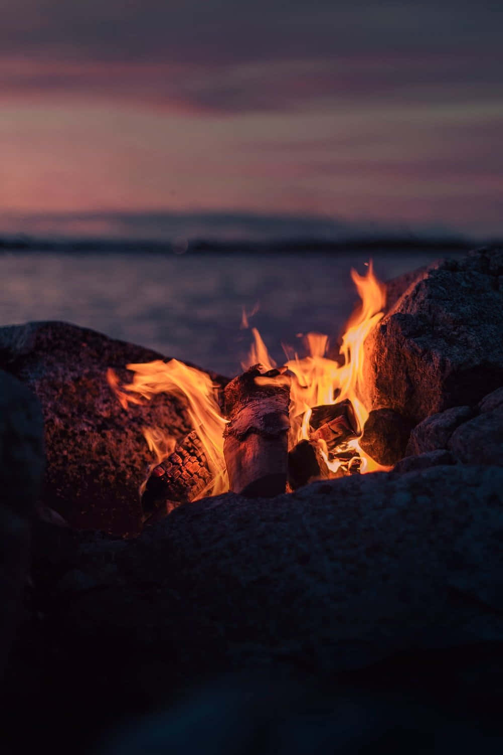 Cozy Fire 桌布 壁紙 — 一個噼啪作響的營火在黑暗的岩石間明亮地燃燒，旁邊是一個平靜的湖泊，上方是塗上粉紅色和紫色色調的暮色天空。