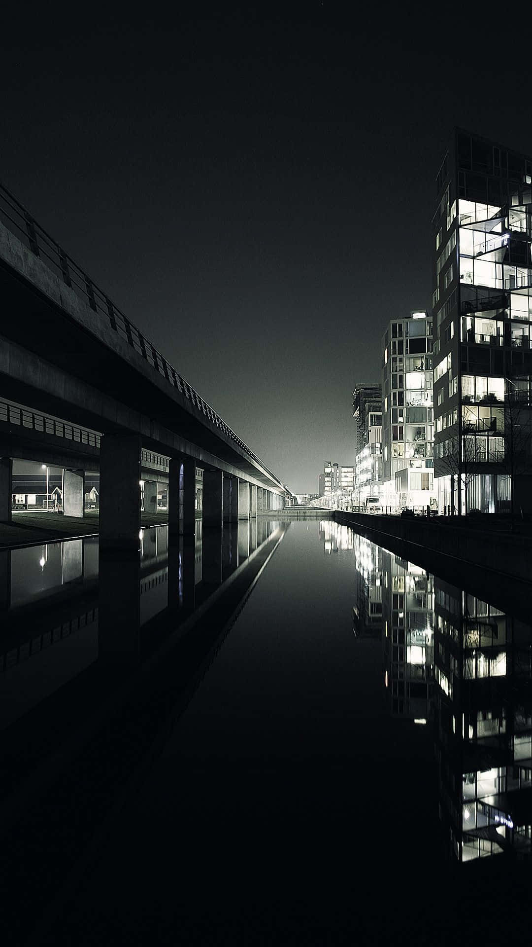 暗夜城市 桌布 壁紙 — 一個嚴峻的夜景城市景觀，發光的窗戶倒映在混凝土天橋下的平靜運河中。