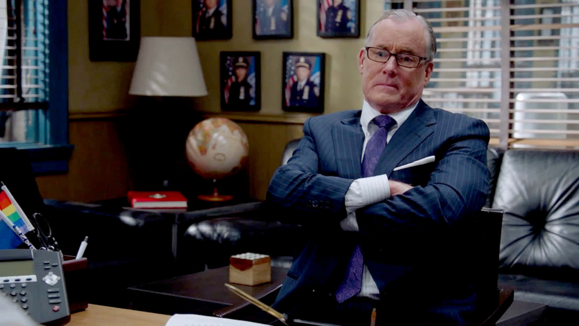 John C. Mcginley 桌布 壁紙 — 一個嚴肅嘅男人著住間條西裝，交叉手臂坐喺皮椅上，喺一個燈光昏暗嘅辦公室，周圍有裱框相片同一個地球儀。