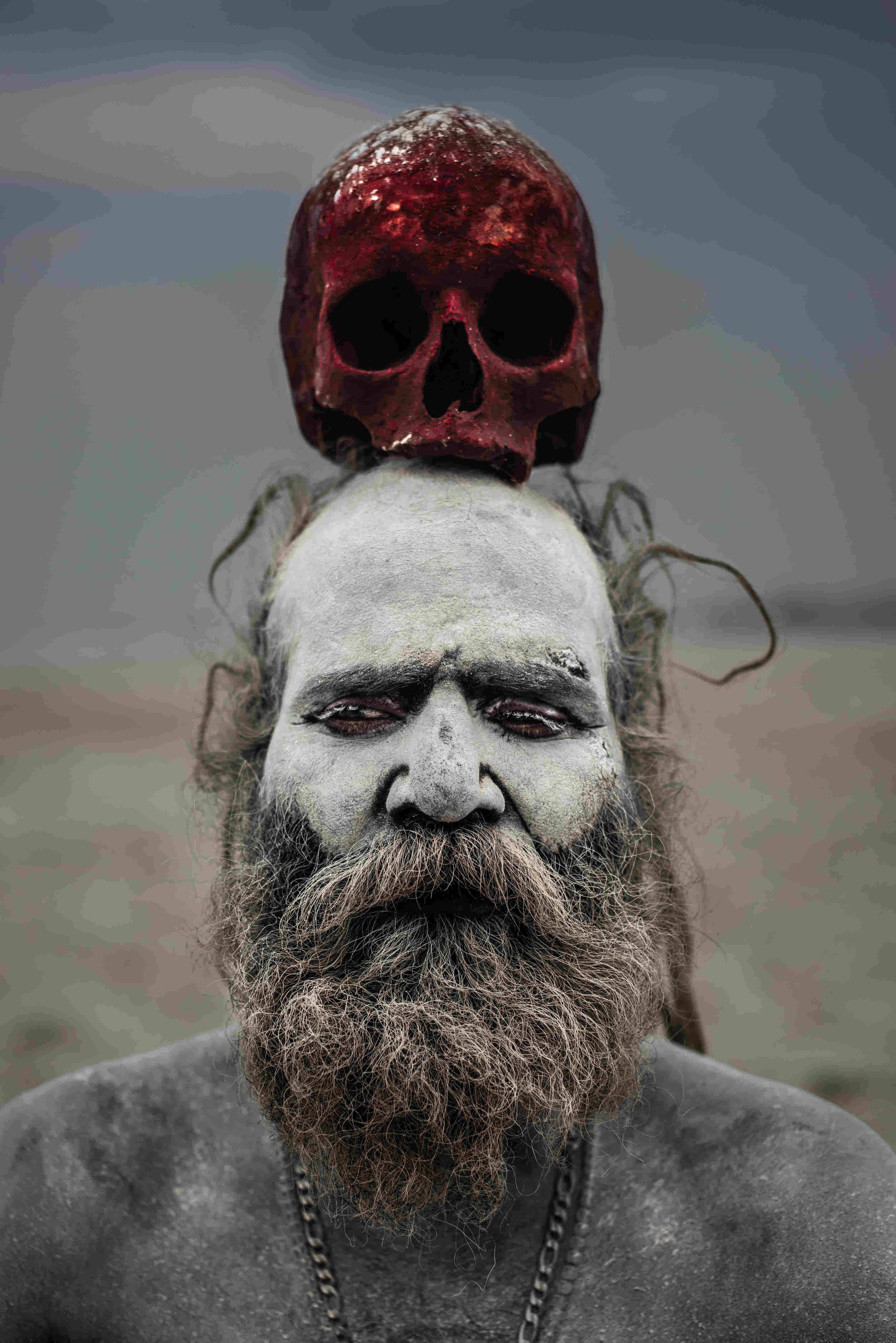 Aghori 桌布 壁紙 — 一個嚴肅的男人，臉上和鬍子塗成白色，在暗淡的天空下將一個紅色頭骨平衡在頭頂上。