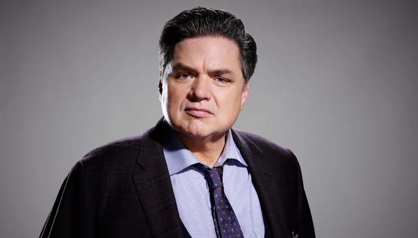 Oliver Platt 桌布 壁紙 — 一個嚴肅的男人穿著深色西裝和圖案領帶，在純灰色工作室背景前直視鏡頭。