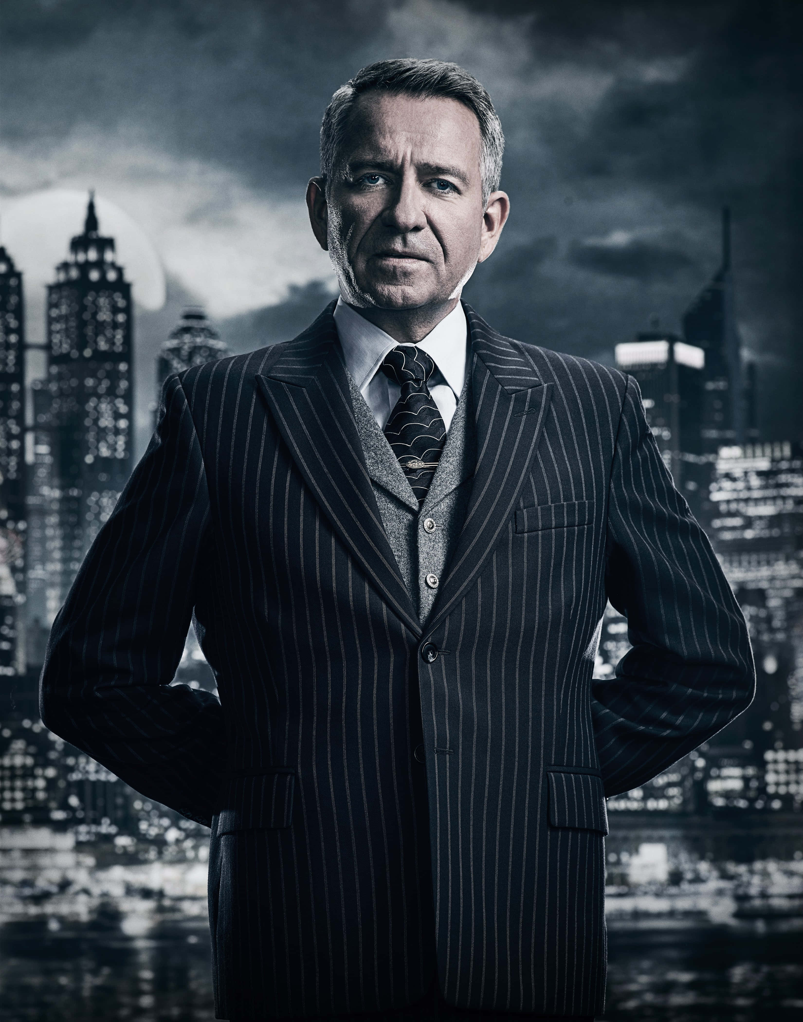 Alfred Pennyworth 桌布 壁紙 — 一個嚴肅的男人穿著細條紋西裝，在夜晚充滿氛圍、燈火通明的城市天際線前自信地站立。