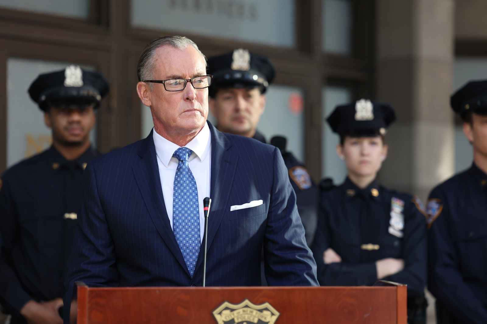 John C. Mcginley 桌布 壁紙 — 一個嚴肅的男人穿著西裝和眼鏡在木製講台上發言，身後站著穿制服的警察。