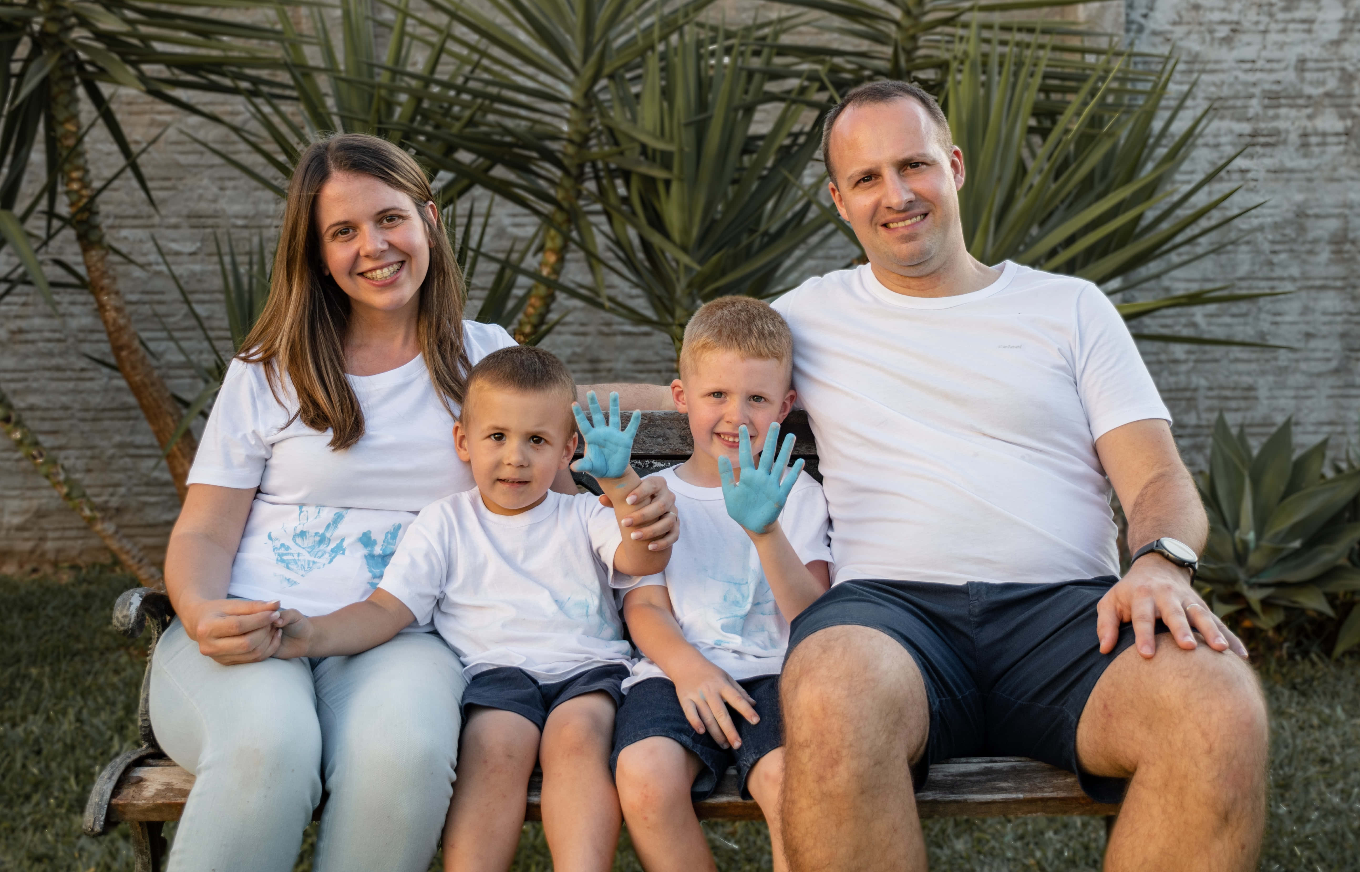 Family Portrait 桌布 壁紙 — 一個四口之家面帶微笑，坐在戶外的木長椅上，手上和襯衫上沾有藍色油漆，背景是棕櫚植物和石牆。
