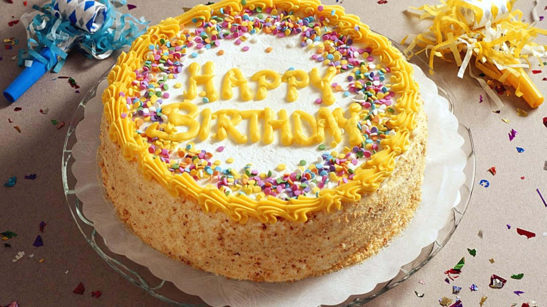 生日蛋糕 桌布 壁紙 — 一個圓形生日蛋糕，上面有黃色糖霜和彩色糖粒，放在玻璃碟上，周圍有五彩紙屑和派對吹龍。