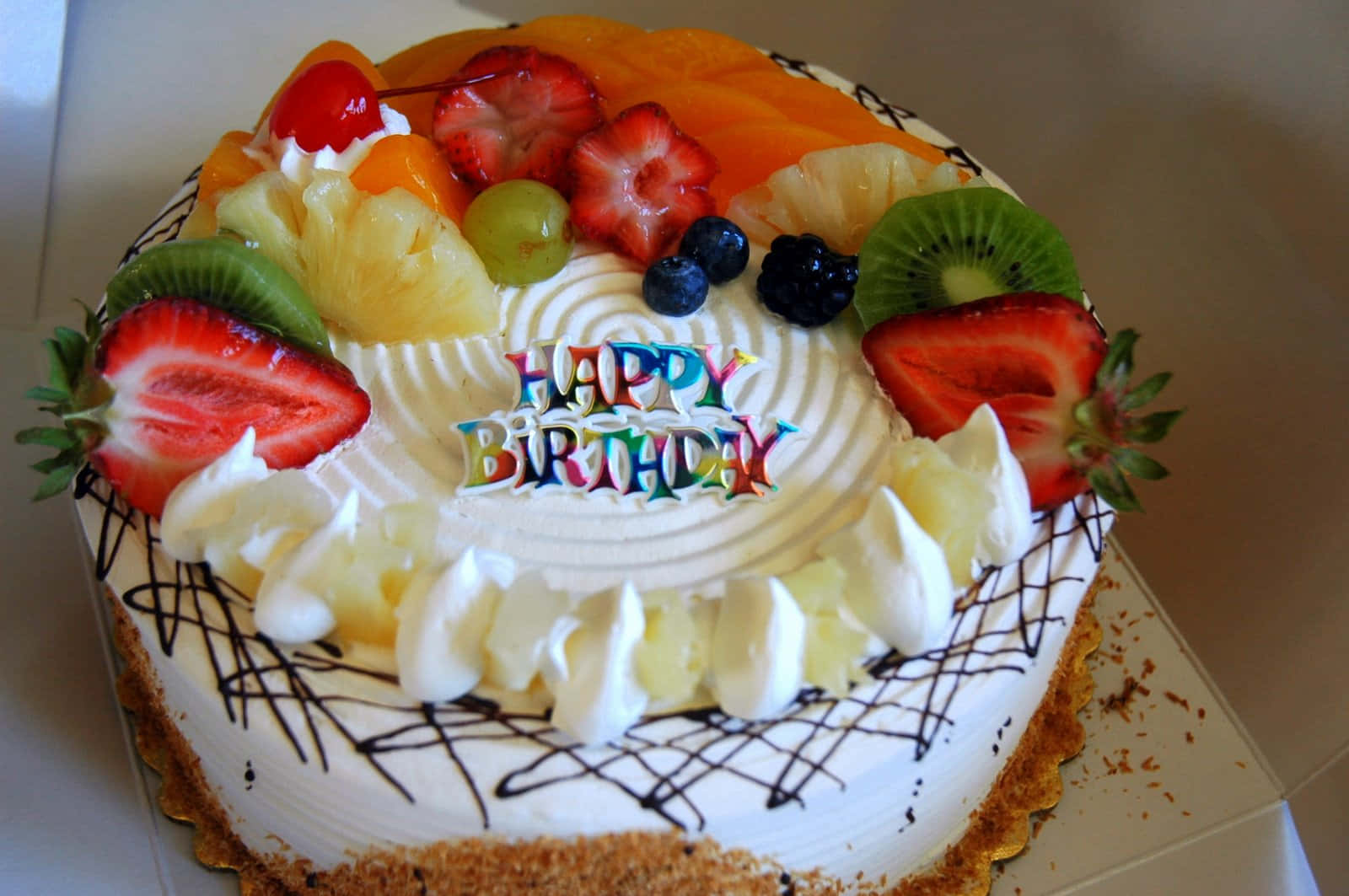 生日蛋糕 桌布 壁紙 — 一個圓形生日蛋糕，上面鋪滿色彩繽紛的新鮮水果，中間插著一支「Happy Birthday」蠟燭。
