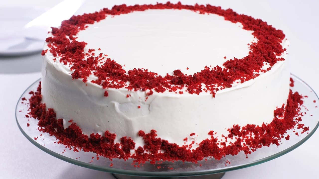 Red Velvet Cake 桌布 壁紙 — 一個圓形的紅絲絨蛋糕，配上白色糖霜和紅色碎屑，擺放在玻璃蛋糕架上，背景是潔淨的白色。