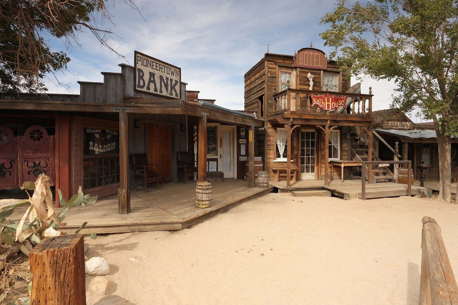 Wild West 桌布 壁紙 — 一個塵土飛揚的西部小鎮廣場，木製建築物上標有銀行和酒店，在明亮的藍天下。