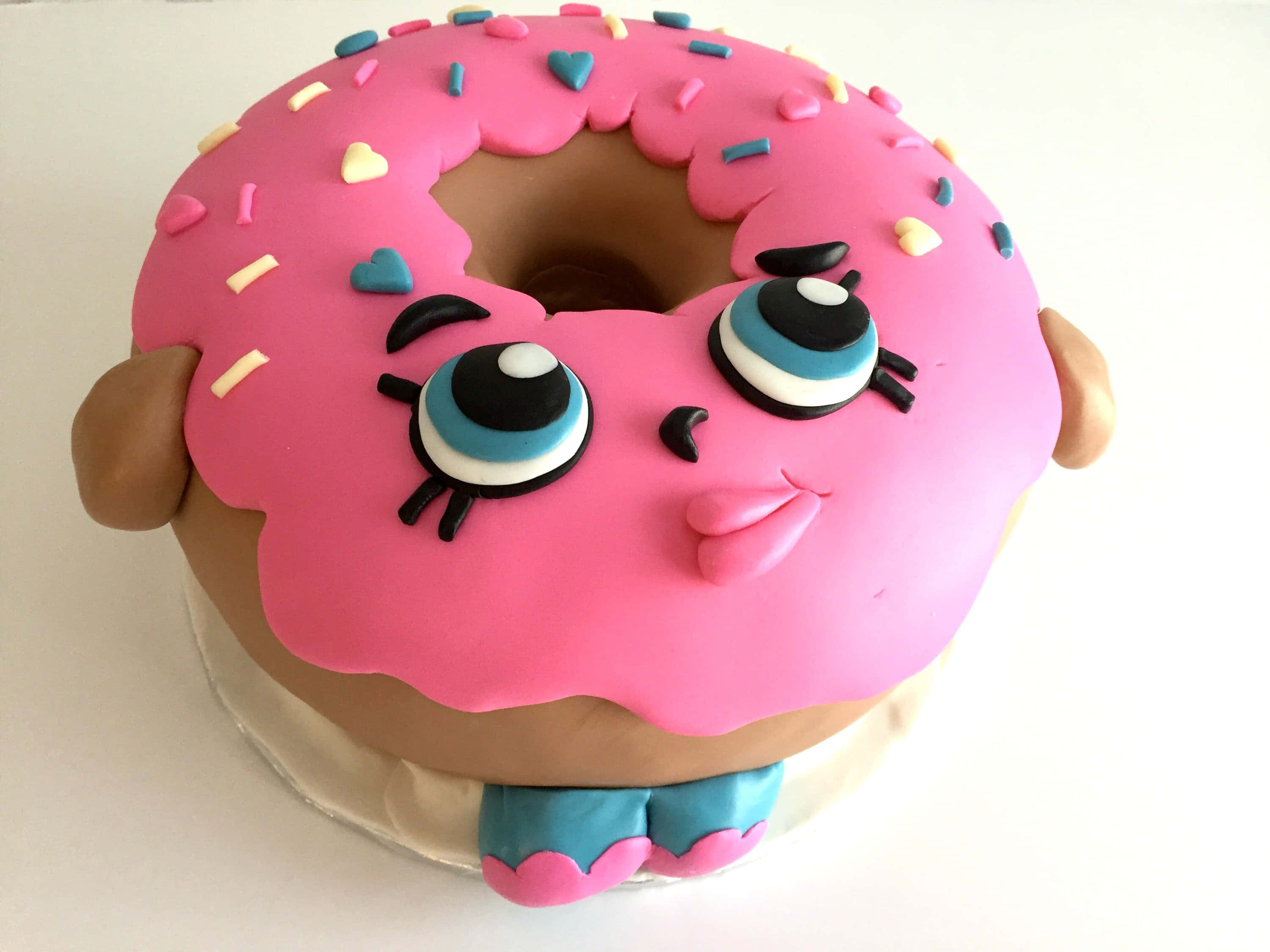 Shopkins 桌布 壁紙 — 一個夢幻的粉紅色糖霜甜甜圈蛋糕，帶有卡通眼睛和心形糖粒，在白色背景上看起來很開心。