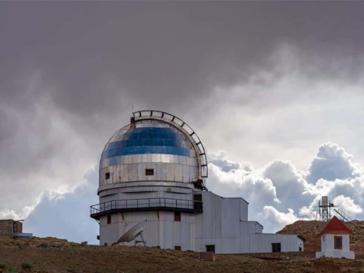 Observatory 桌布 壁紙 — 一個大型天文台圓頂，帶有藍色和銀色面板，矗立在荒涼的山丘上，天空多雲，氣氛戲劇化。