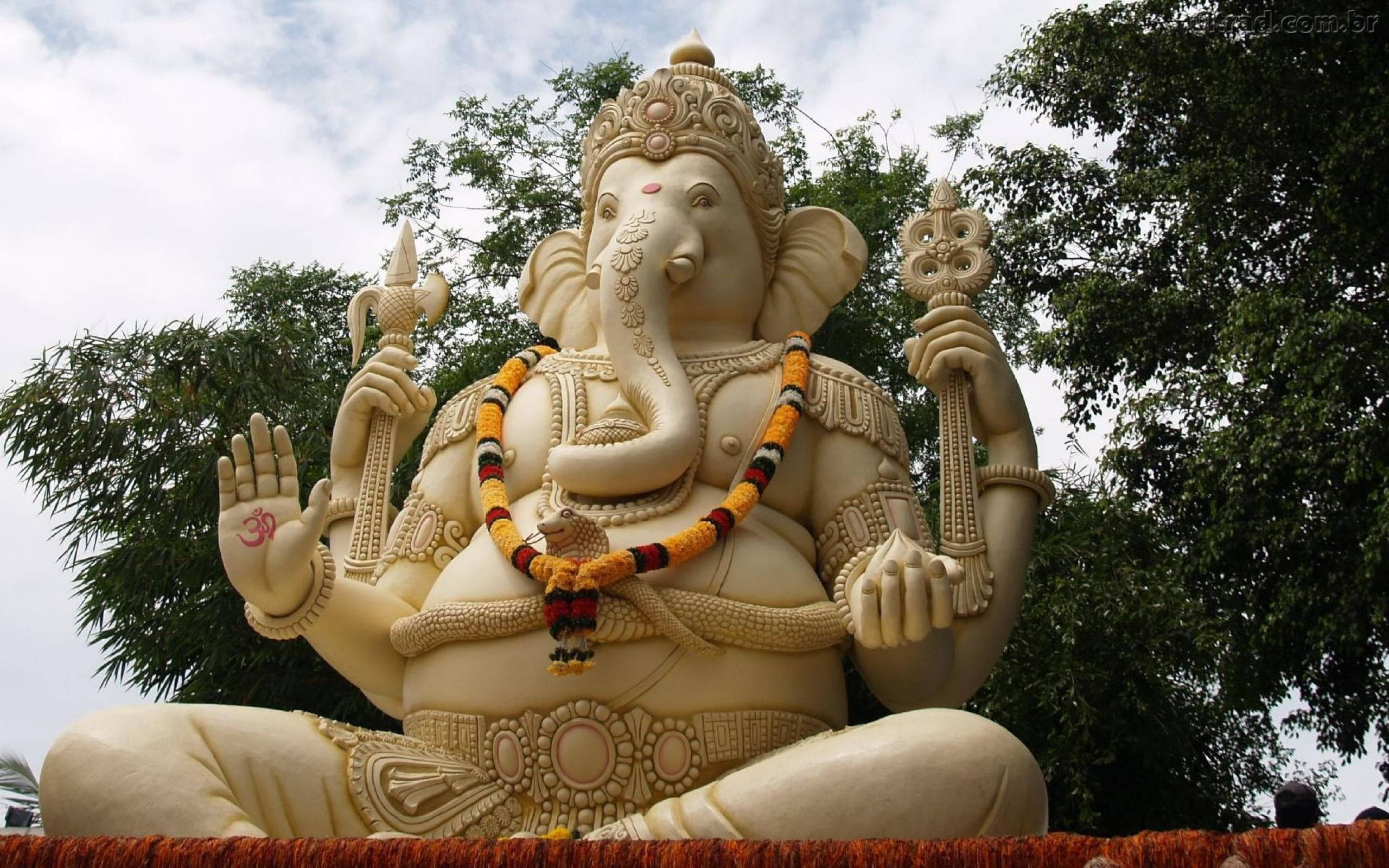 Ganesh Chaturthi 桌布 壁紙 — 一個大型奶油色象神雕像，飾以色彩繽紛的花環，在部分多雲的天空下寧靜地坐於戶外。