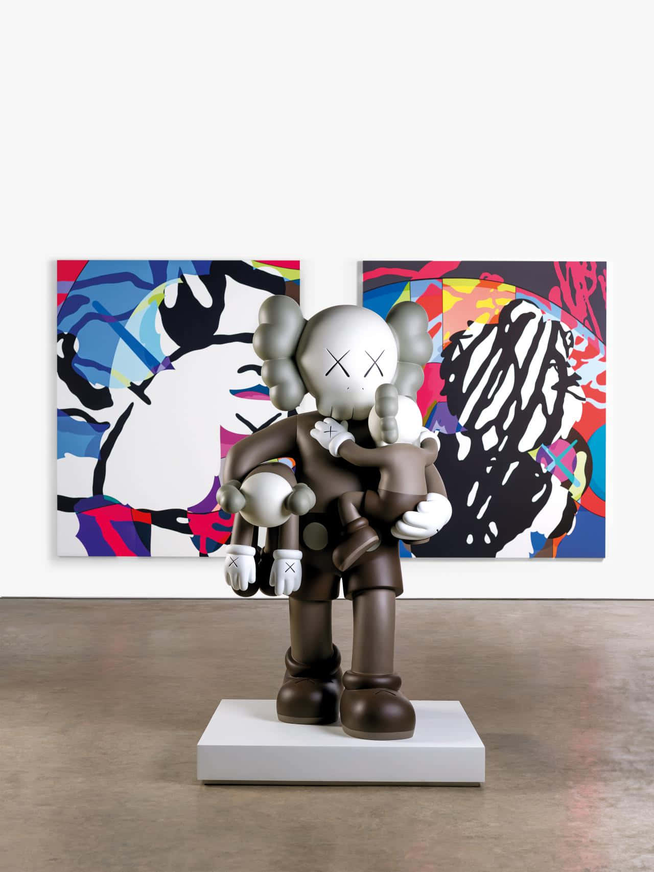 Kaws Figures 桌布 壁紙 — 一個大型棕色KAWS雕塑手持一個較小的雕像，站在白色基座上，前面是兩幅色彩繽紛的抽象畫。