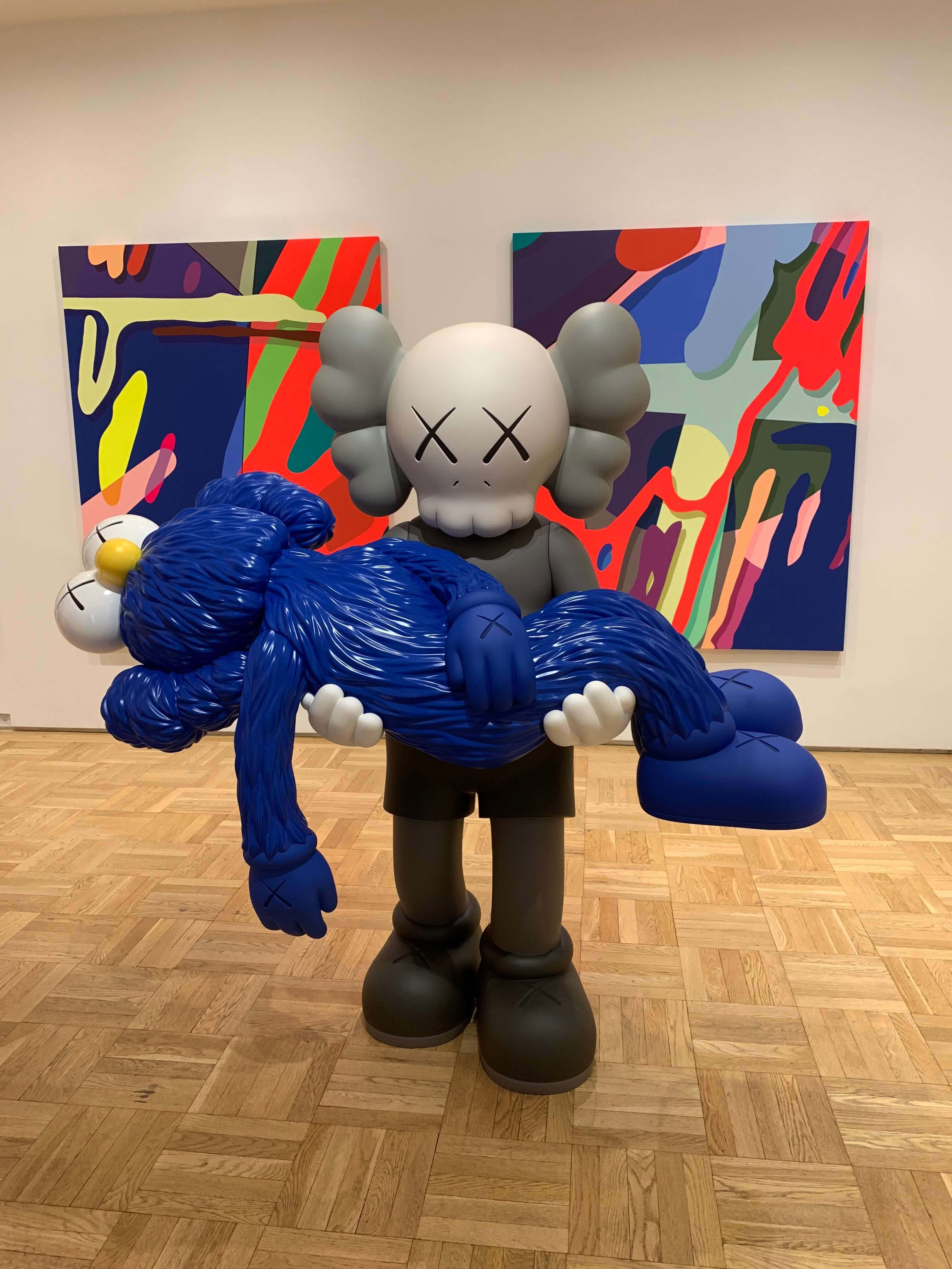 Kaws Bff 桌布 壁紙 — 一個大型灰色KAWS雕塑在畫廊中抱著一個藍色Companion公仔，牆上掛滿色彩繽紛的抽象畫。
