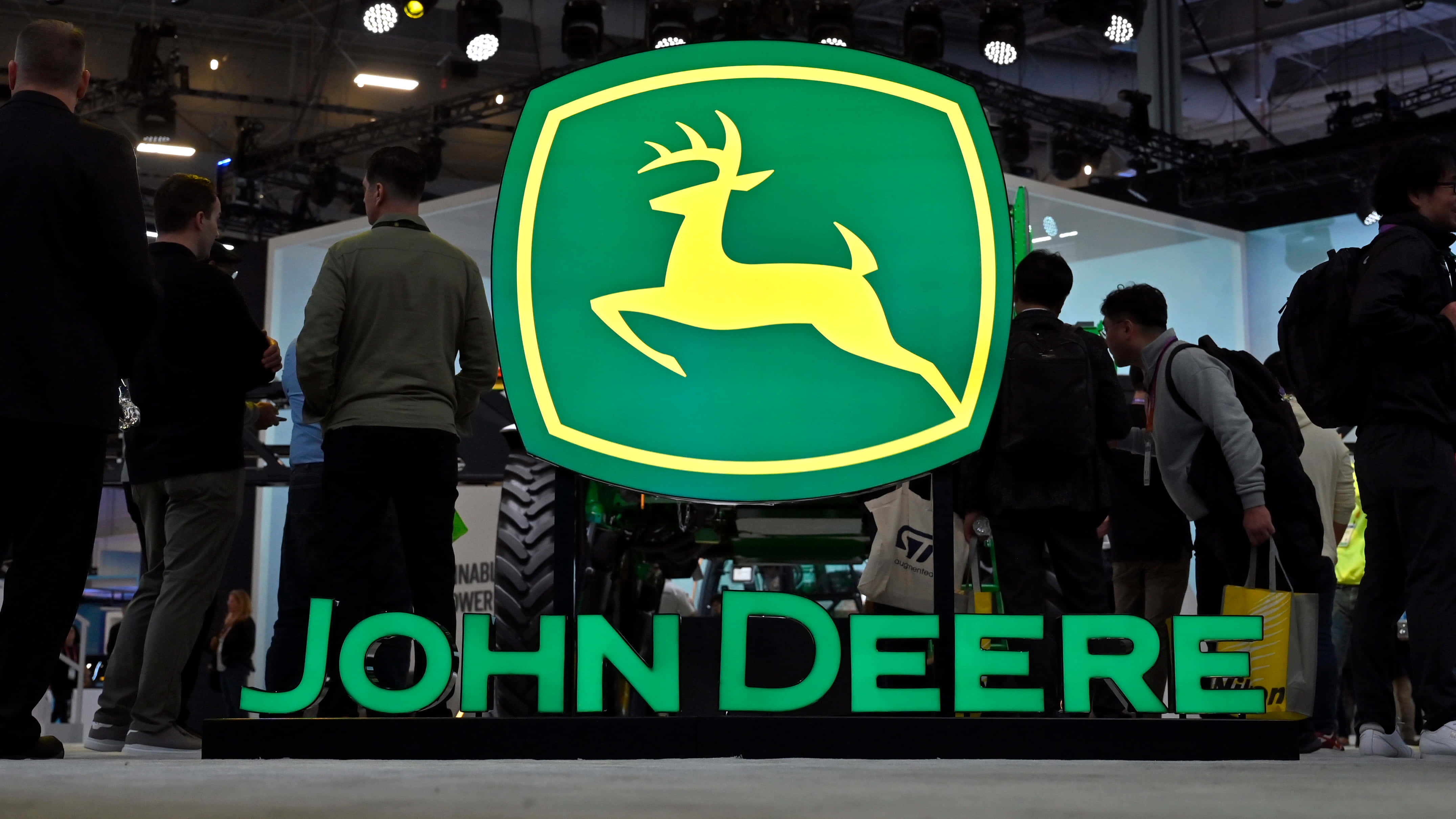 John Deere 桌布 壁紙 — 一個大型發光的John Deere標誌，帶有一隻跳躍的鹿，在繁忙的室內展覽廳的貿易展上醒目地矗立，周圍有與會者。