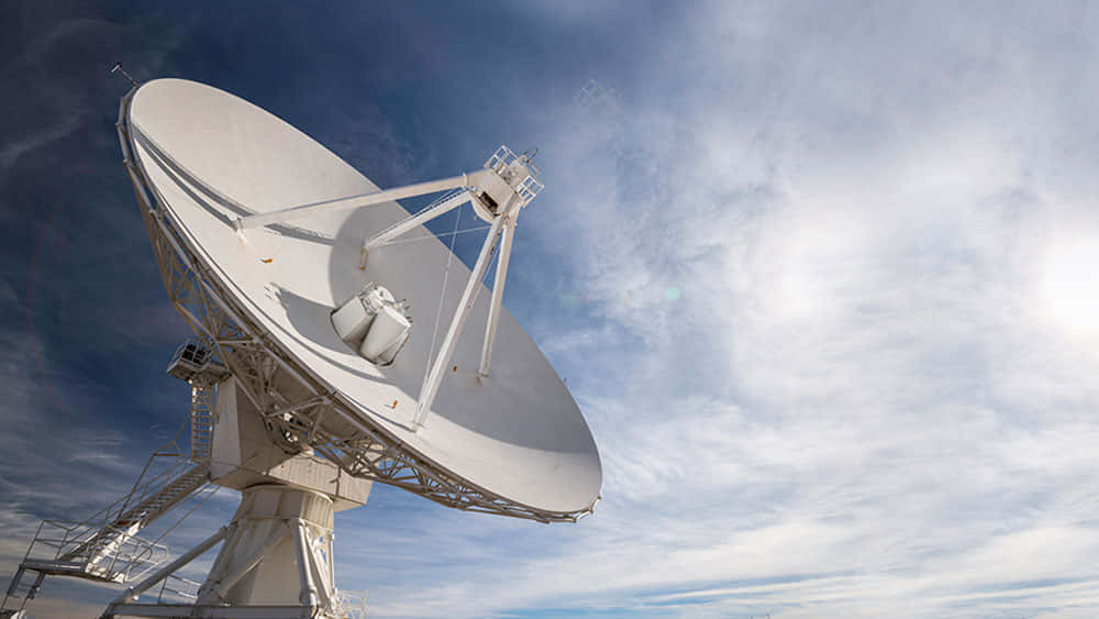 Satellite Technology 桌布 壁紙 — 一個大型白色衛星天線向上傾斜，背景是帶有薄雲和陽光眩光的明亮藍天。
