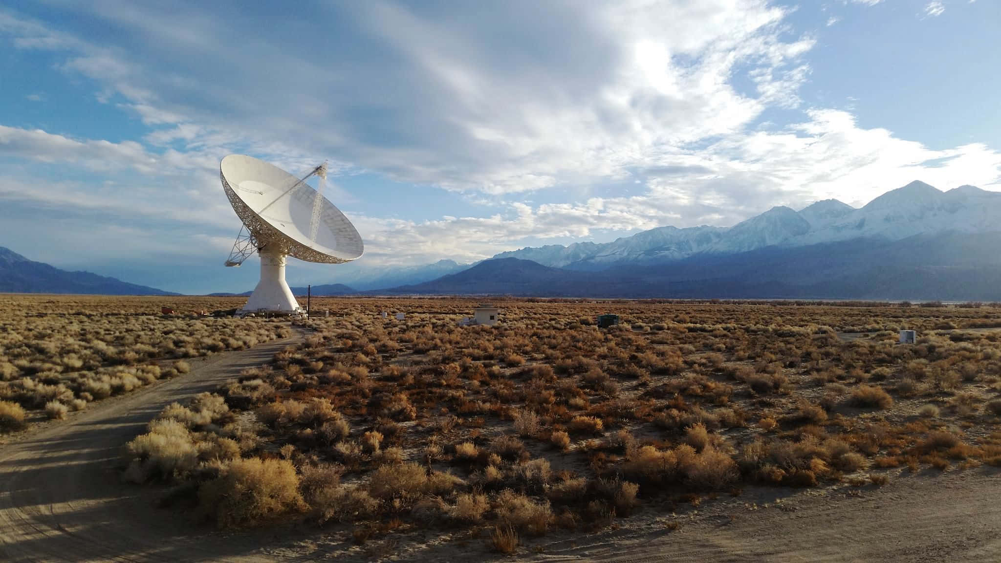 Radio Telescope 桌布 壁紙 — 一個大型白色衛星天線獨自矗立在乾燥的沙漠景觀中，遠處有積雪覆蓋的山脈，天空部分多雲。