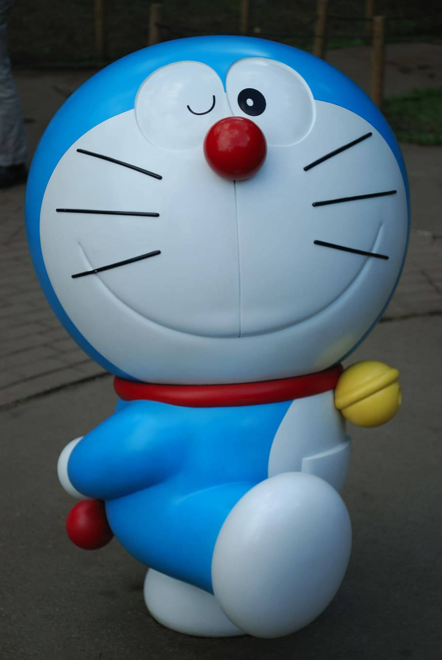 Doraemon 桌布 壁紙 — 一個大型藍色機械貓雕像，有紅色鼻子和黃色鈴鐺，微笑地坐在鋪砌的小路上。