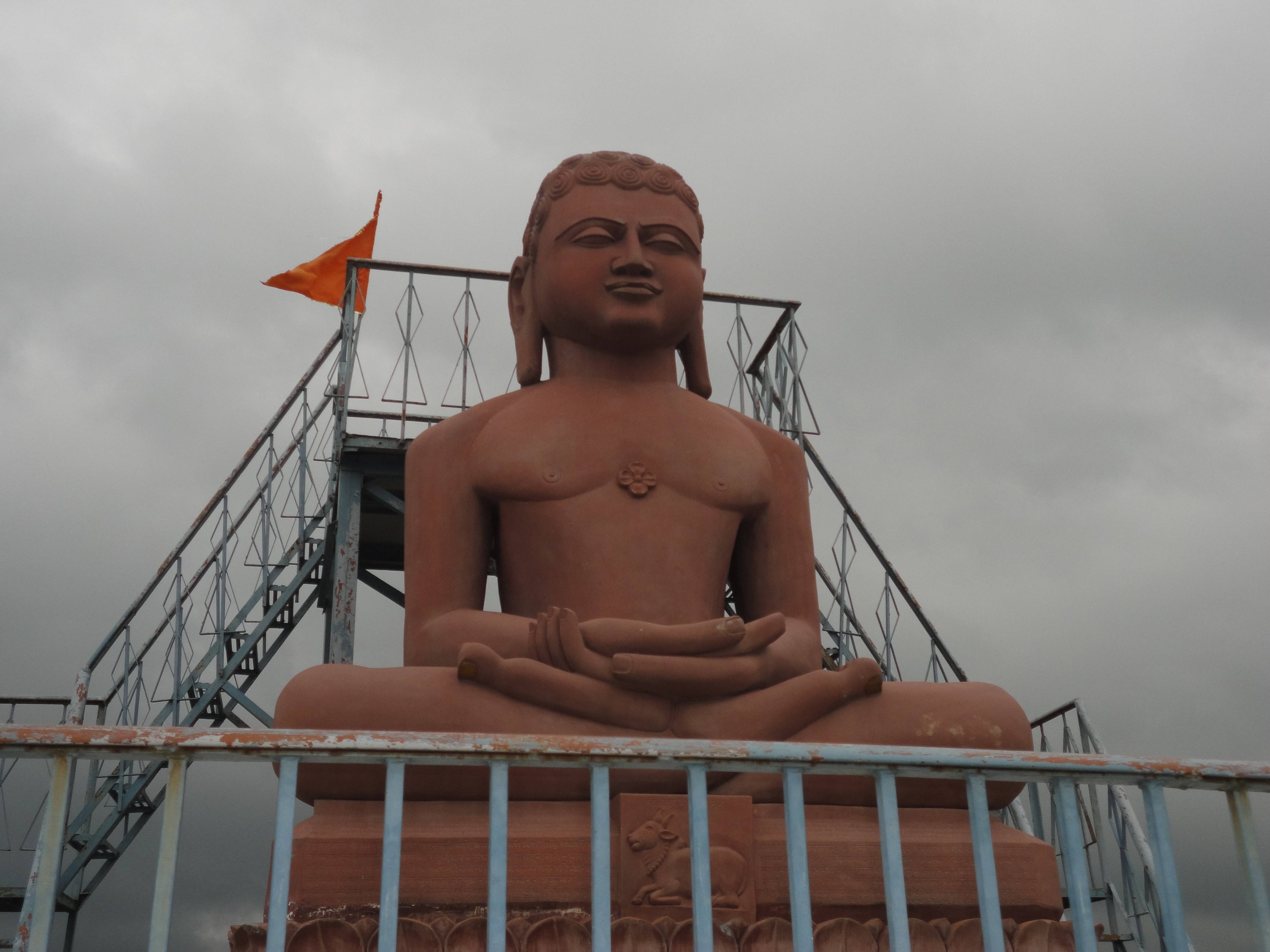 Buddha 桌布 壁紙 — 一個大型赤陶佛像在陰天底下，安詳地坐在金屬平台上冥想，附近有一面橙色旗幟飄揚。