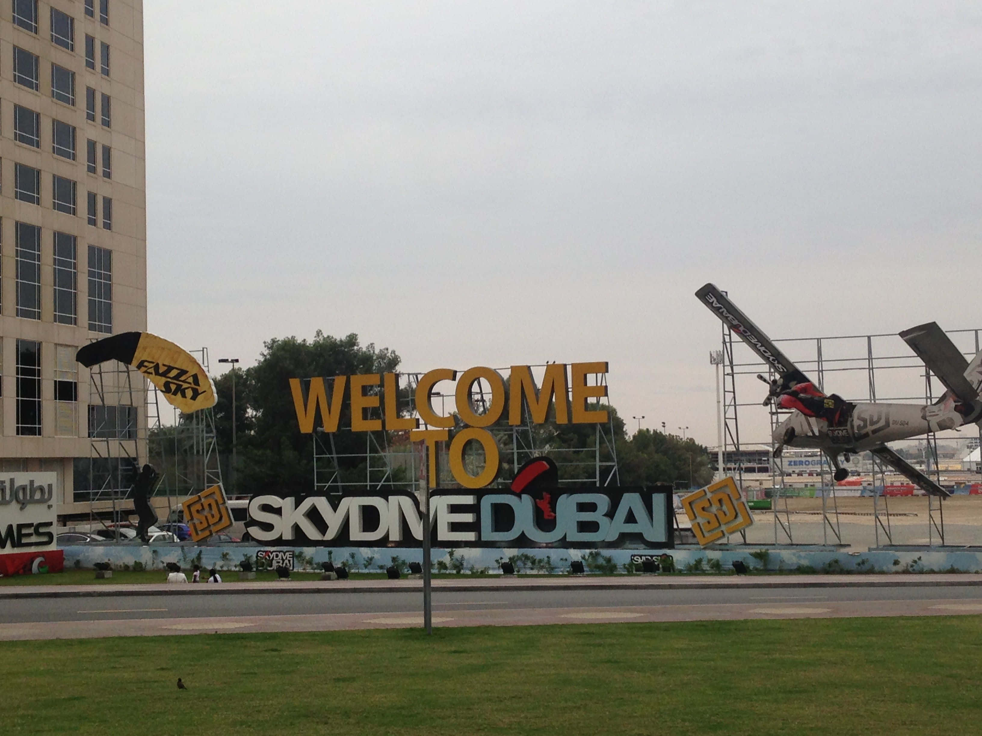 Dubai 桌布 壁紙 — 一個大型黃色標牌，上面寫著「WELCOME TO SKYDIVE DUBAI」，旁邊懸掛著一架飛機和一個降落傘，背景是多雲的天空。