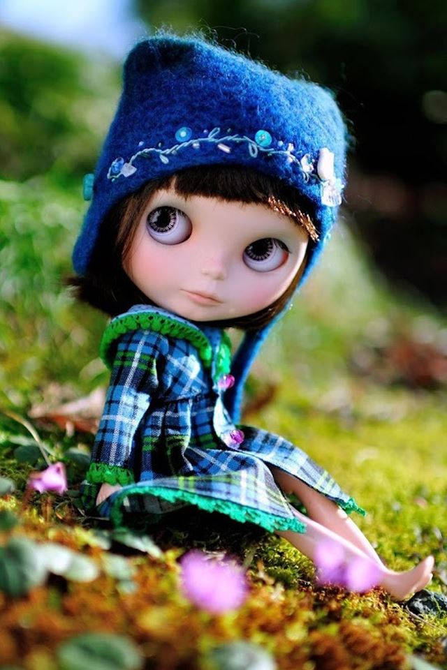Doll 桌布 壁紙 — 一個大眼睛的洋娃娃戴著藍色帽子和格子裙，坐在長滿苔蘚的地上，周圍有紫色花朵。