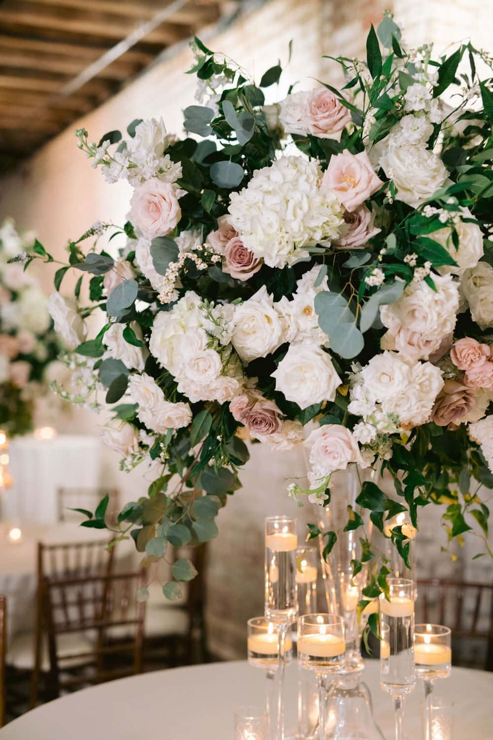 Roses Wedding 桌布 壁紙 — 一個奢華的花卉中心裝飾，配有白色和淡粉紅玫瑰、尤加利葉和蠟燭，在鄉村婚禮場地的桌子上溫暖地閃耀。