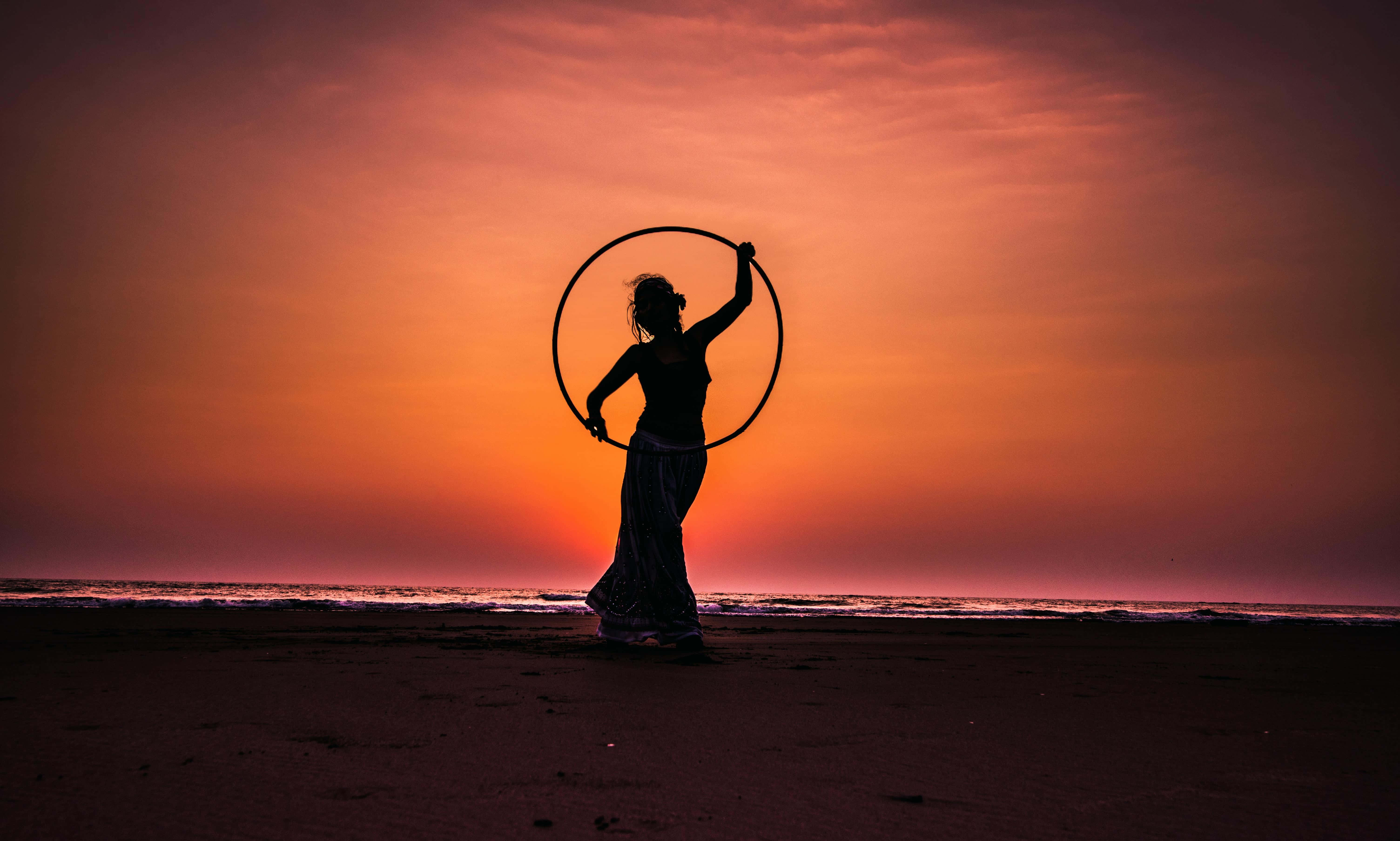 Sunset Silhouette 桌布 壁紙 — 一個女人優雅地拿著呼拉圈的剪影，背景是海洋上熾熱的橙色和粉紅色日落。