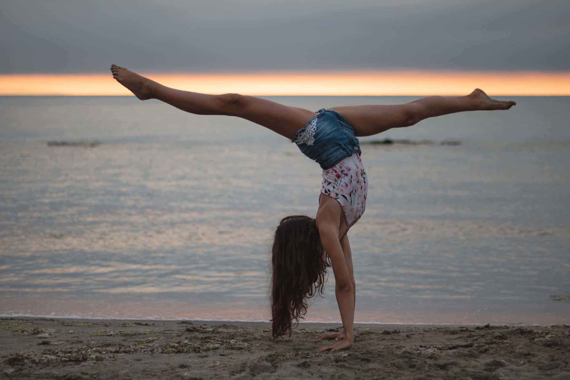 Gymnastics 桌布 壁紙 — 一個女孩在日落時分的沙灘上做倒立劈腿，身後是平靜的海水。