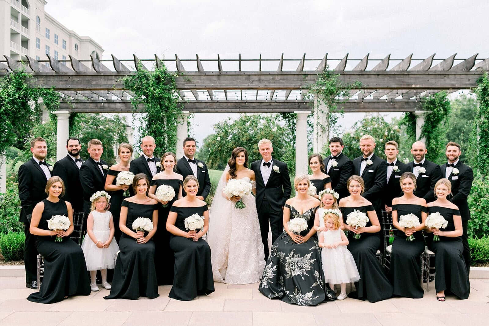 Black And White Wedding 桌布 壁紙 — 一個婚禮派對在藤蔓覆蓋的涼亭下擺姿勢，新娘身穿白色婚紗，新郎身穿燕尾服，周圍是身穿黑色連衣裙的伴娘和身穿黑色燕尾服的伴郎，手持白色花束。